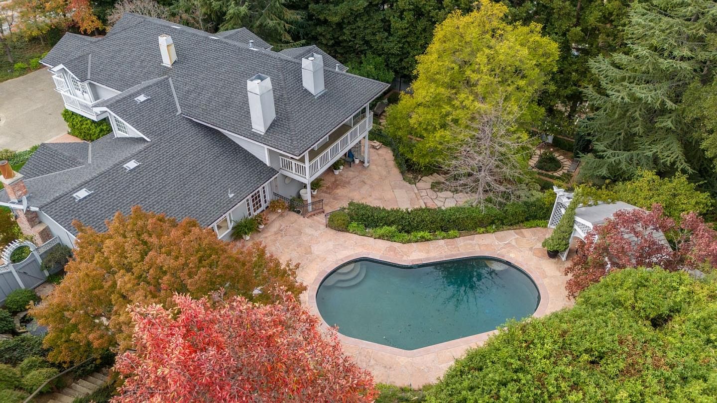 This aerial shot showcases a luxurious home with a dark gray roof, white exterior, and multiple chimneys. The backyard features a heart-shaped pool surrounded by a stone patio and lush landscaping, including mature trees with vibrant fall colors. A charming white gazebo and a meticulously maintained garden add to the property's appeal.