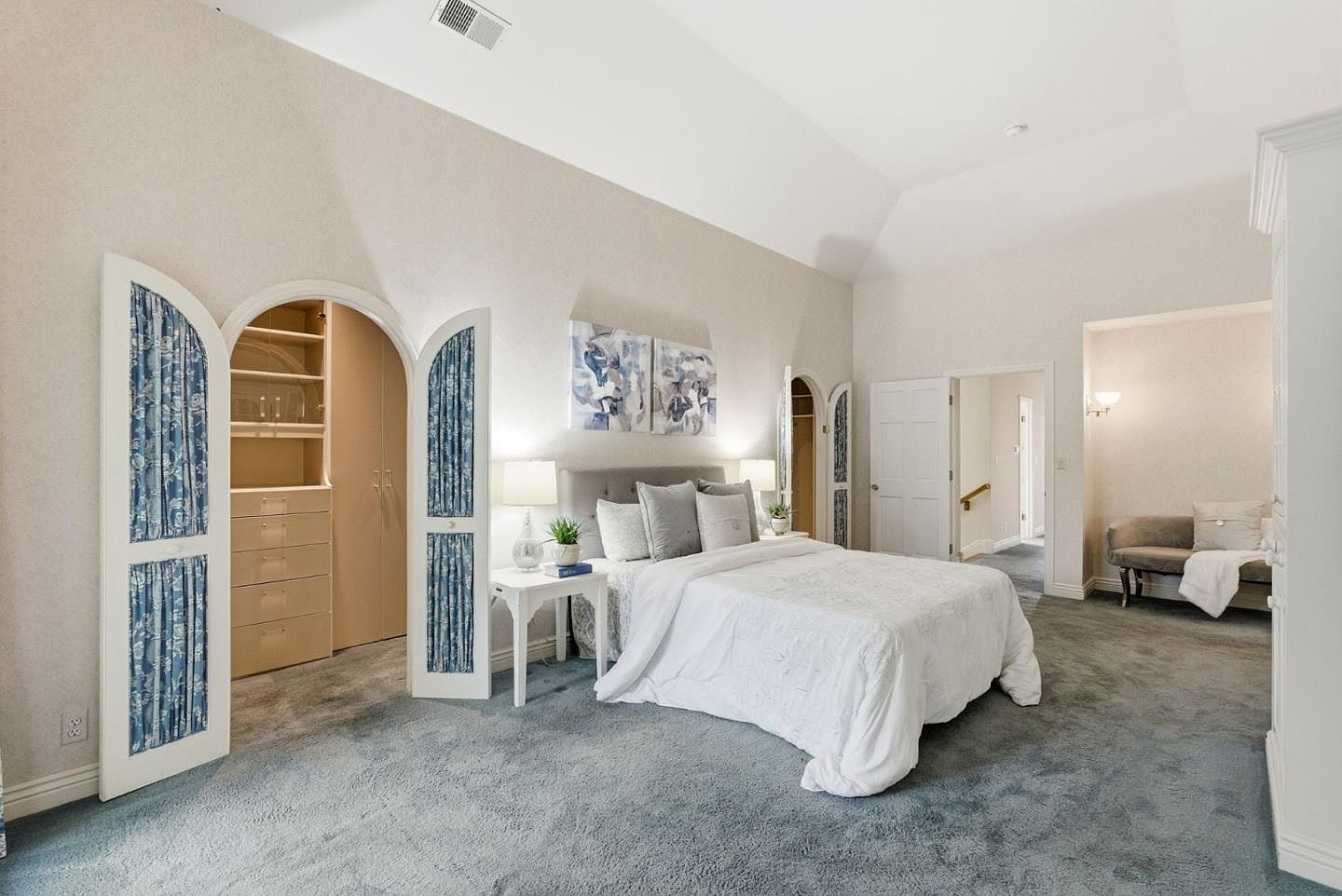 This is a primary bedroom featuring a large bed with white bedding and gray pillows, flanked by matching nightstands and lamps. The room has an ensuite walk-in closet with arched doors and blue patterned fabric, and a seating area is visible through a doorway. The overall impression is spacious and serene, with a soft color palette and plush carpeting.
