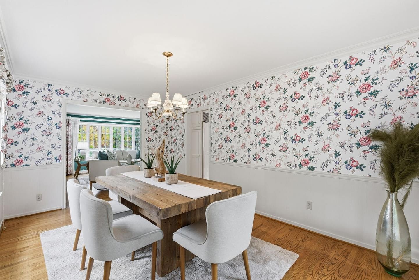 This is an interior shot of a dining room featuring a rustic wooden table surrounded by upholstered chairs. The walls are adorned with floral wallpaper above white wainscoting, and a chandelier hangs above the table. The room has a bright and airy feel, enhanced by natural light and a view into an adjacent living area.