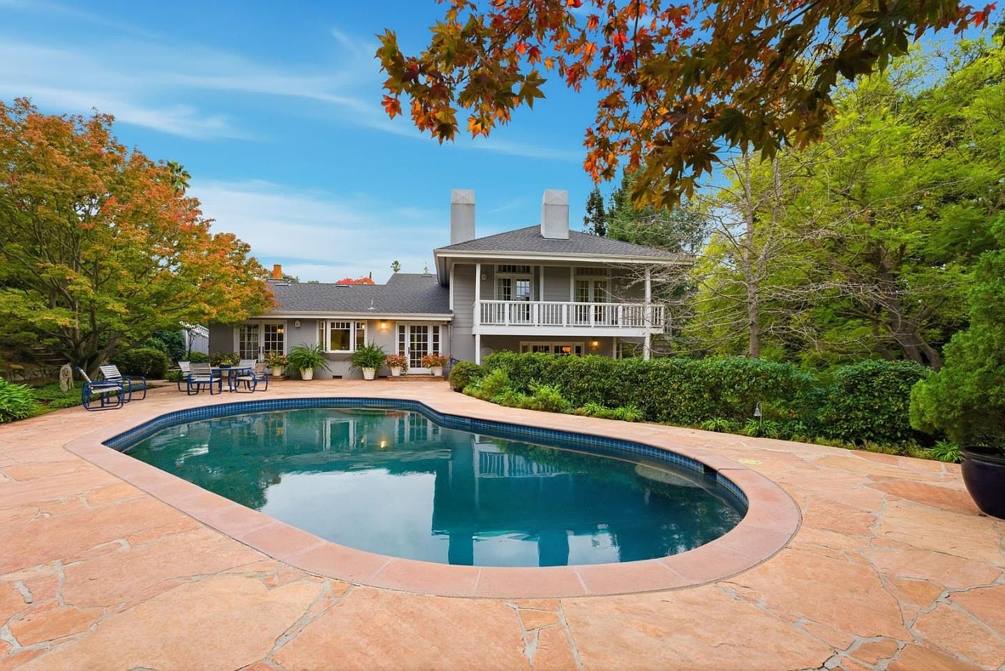 This image showcases a luxurious backyard featuring a kidney-shaped pool with a stone surround. The pool reflects the house, which has a balcony and multiple windows, and is surrounded by lush greenery and mature trees, creating a private and serene oasis. The scene suggests a high-end property perfect for relaxation and outdoor entertaining.