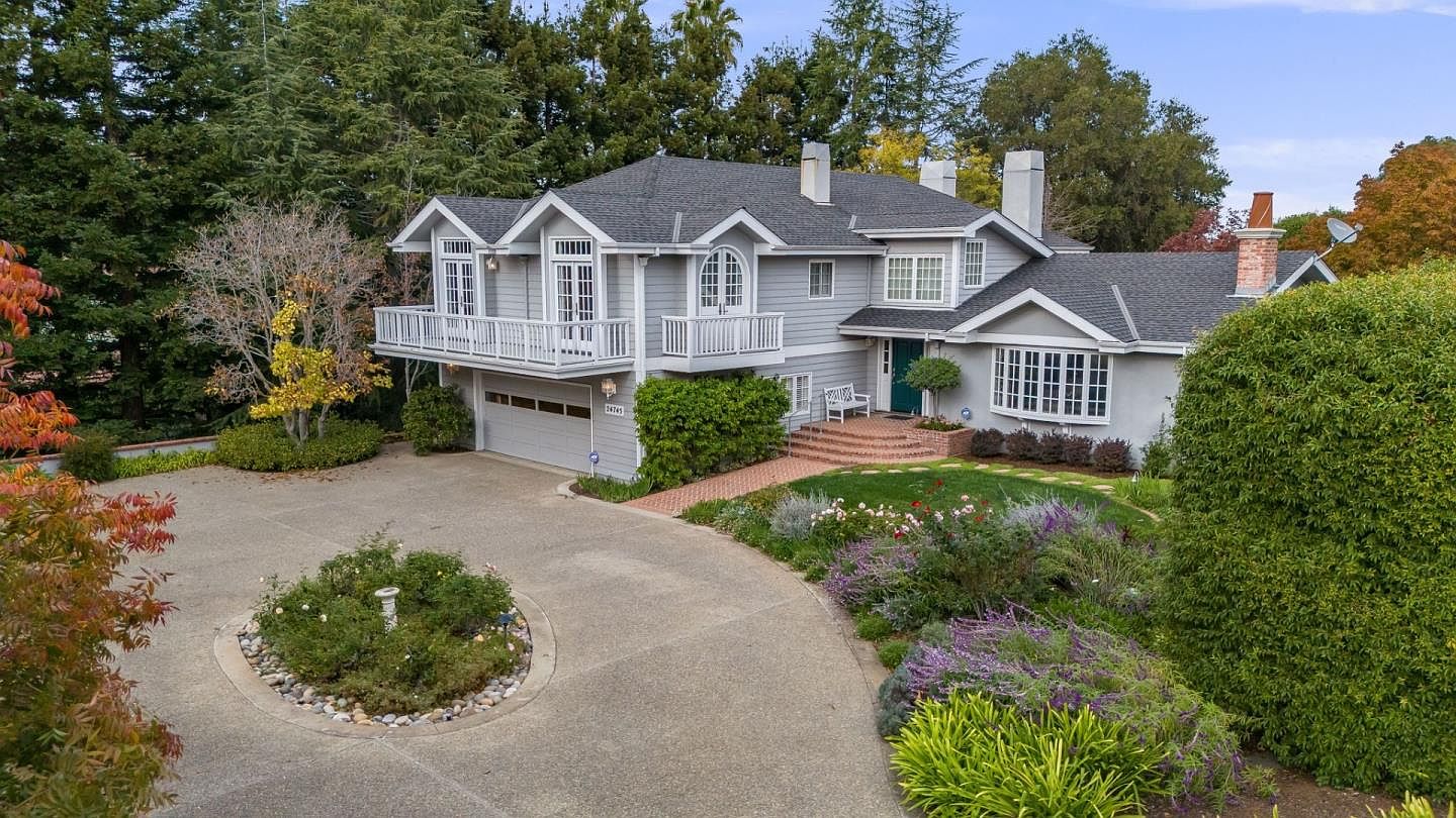 This is an aerial front view of a two-story gray house with a dark gray roof, white trim, and a two-car garage. The house features a balcony above the garage, a bay window, and a green front door. The property is well-landscaped with a circular driveway, mature trees, and colorful flower beds, creating a welcoming and established atmosphere.