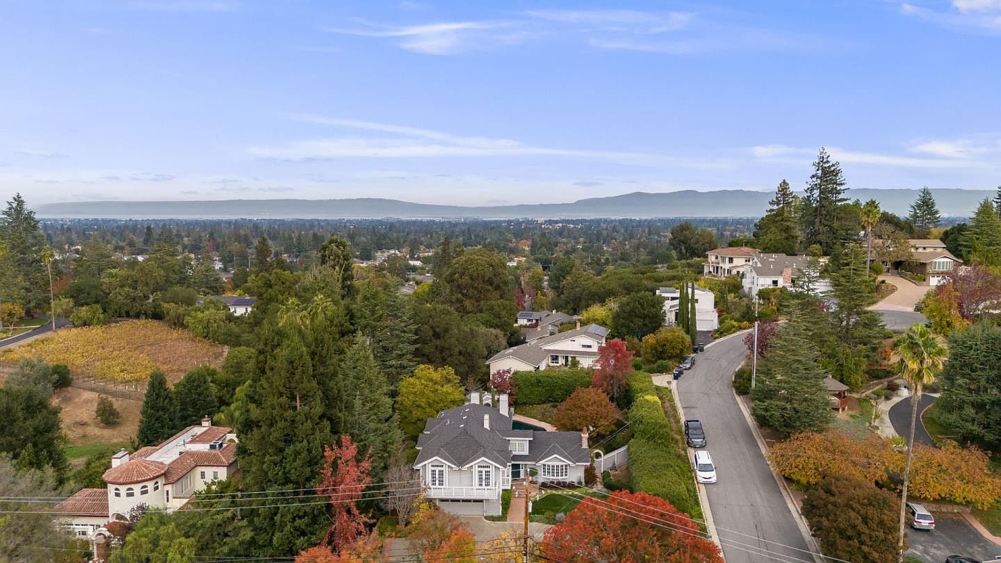 This aerial shot showcases a luxurious residential area with well-maintained homes nestled among lush greenery and mature trees. A winding road leads through the neighborhood, connecting the properties. In the background, a scenic view of distant mountains and a sprawling cityscape adds to the property's appeal, suggesting a desirable location with both natural beauty and urban convenience.