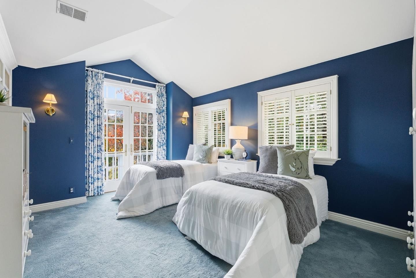 This is a well-lit bedroom featuring two twin beds with white and gray bedding, complemented by gray throw blankets and decorative pillows. The walls are painted a deep blue, contrasting with the white trim and plantation shutters on the windows. Blue and white floral curtains frame the French doors, adding a touch of elegance to the room.