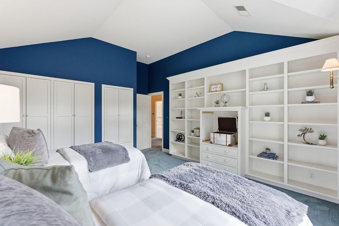 This is a bright and inviting guest bedroom featuring two twin beds with white and gray bedding. The room has a built-in white bookcase and white closets, providing ample storage. The walls are painted a deep blue, creating a cozy and stylish atmosphere.