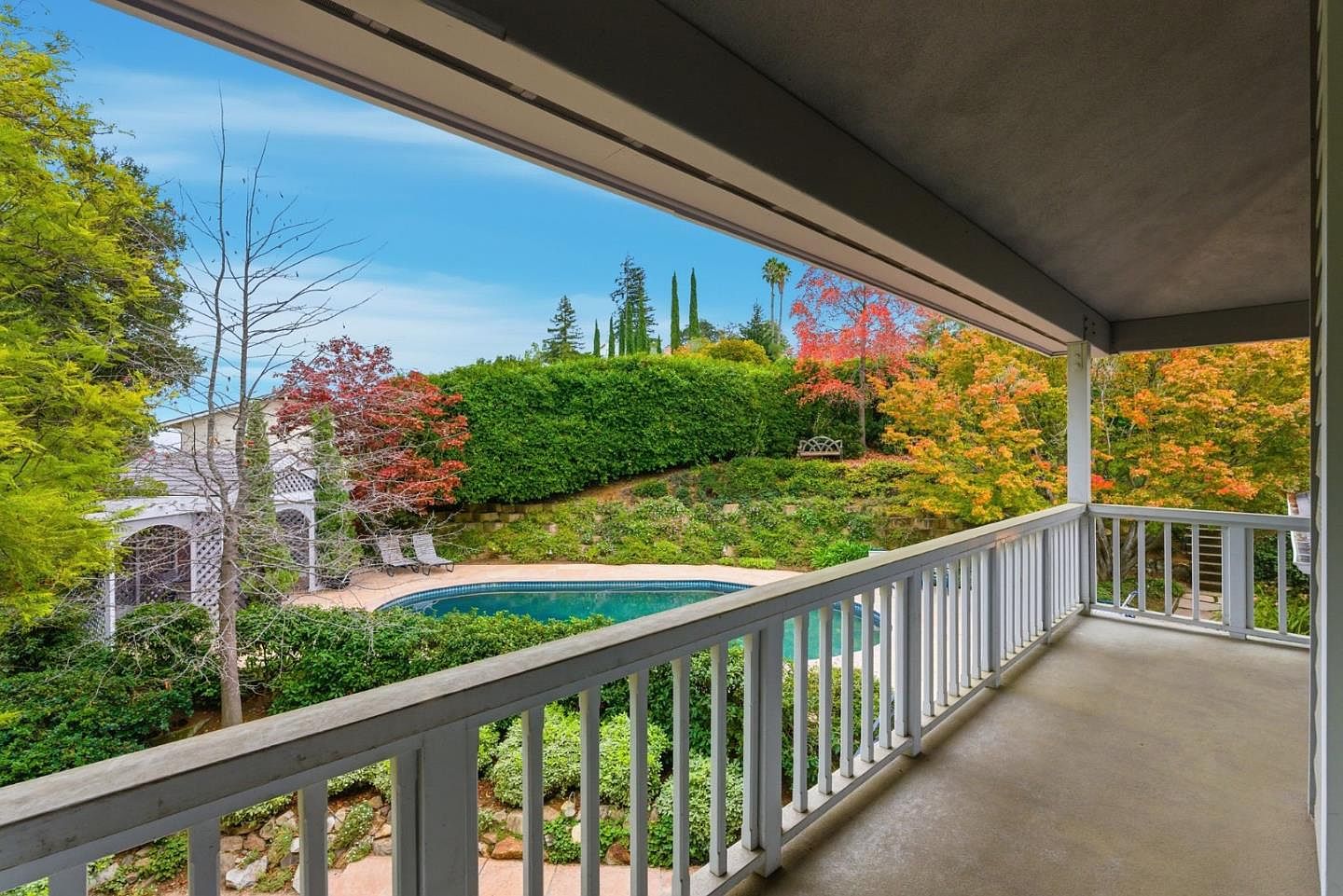 This image showcases a charming balcony view overlooking a lush backyard with a swimming pool. The white railing of the balcony frames the scene, highlighting the well-maintained garden, mature trees with fall colors, and a neatly trimmed hedge. The overall impression is one of tranquility and privacy, perfect for relaxation and outdoor enjoyment.