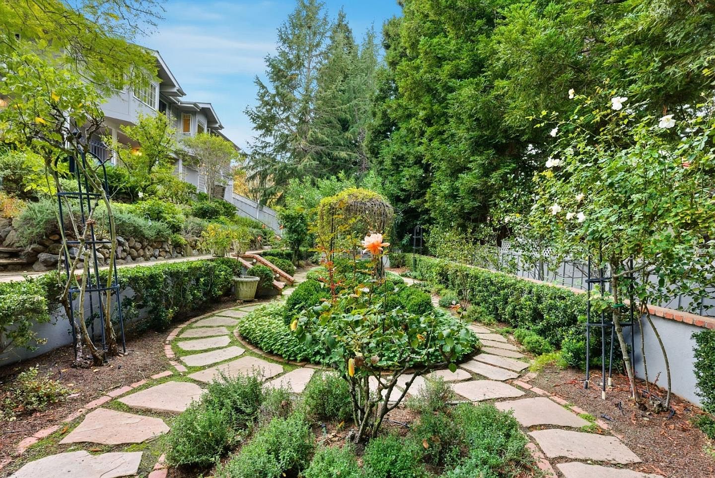 This image showcases a meticulously landscaped garden with a stone pathway winding through lush greenery. A central rose bush with a trellis serves as a focal point, surrounded by manicured hedges and various plants. The garden exudes a sense of tranquility and sophistication, enhancing the property's curb appeal.