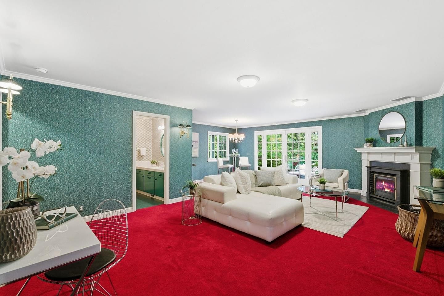 This is an interior shot of a living room featuring a red carpet, teal walls, and white trim. The room includes a white sectional sofa, a fireplace with a white mantel, and a view into a dining area with large windows. The style is somewhat retro with a bold color palette.