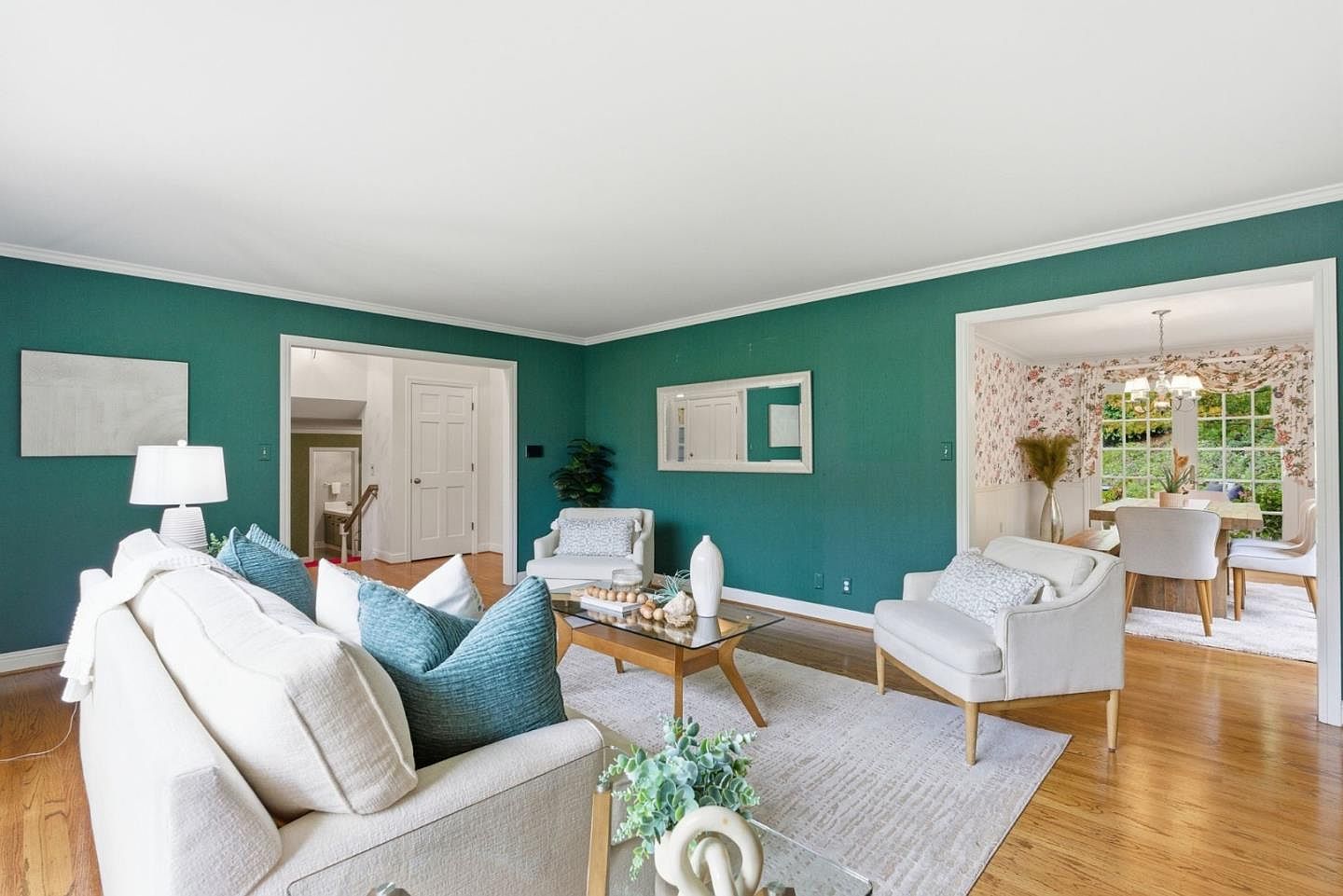 This is an interior shot of a living room featuring a cream-colored sofa with teal pillows, two white armchairs, and a glass-topped coffee table. The walls are painted a deep teal color, complemented by white trim and a light-colored area rug. The room has hardwood floors and an open doorway leading to a dining area, creating a bright and inviting space.