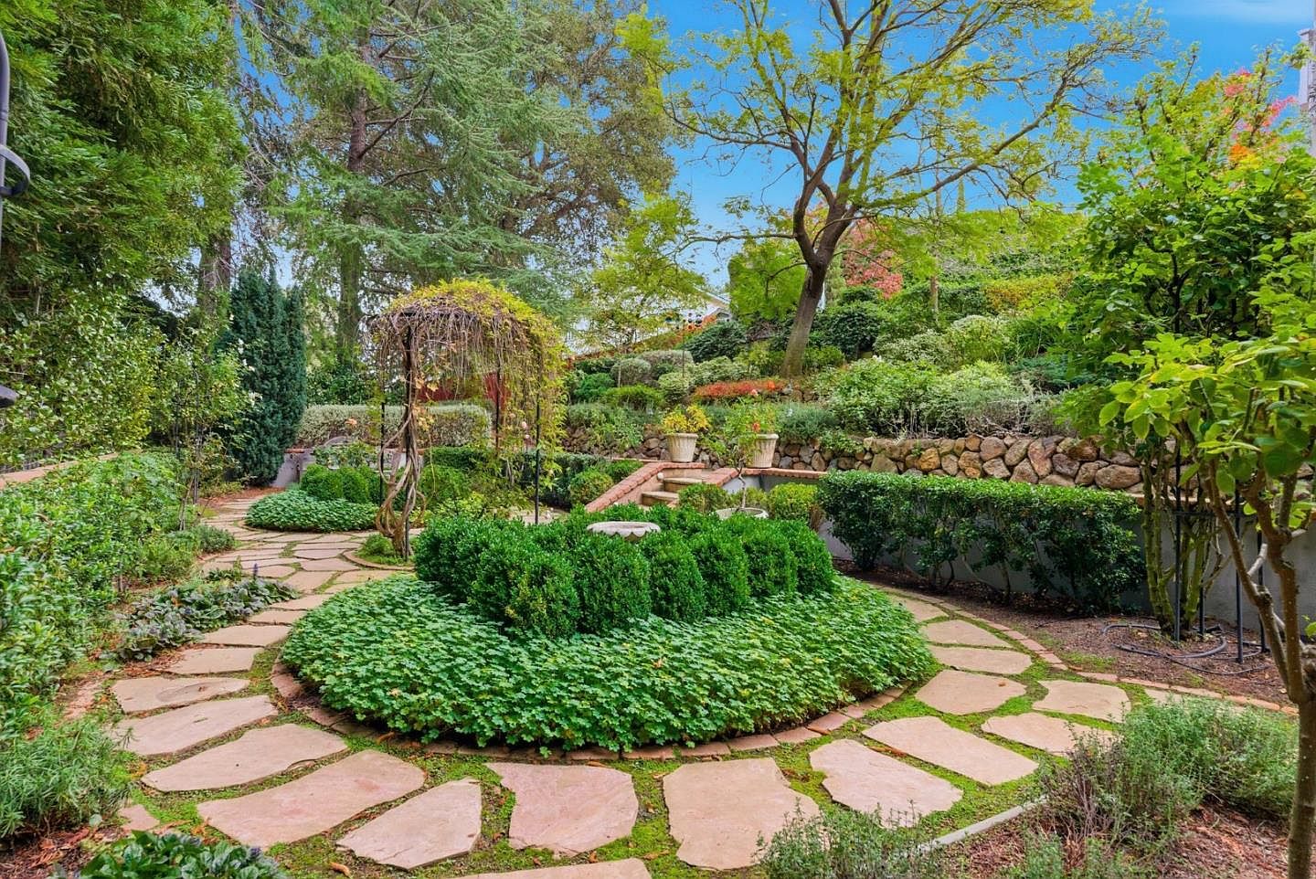 This image showcases a meticulously landscaped garden with a variety of plants, stone pathways, and a charming arbor. The garden features a circular bed of lush greenery surrounded by stone pavers, leading to different areas of the yard. The overall impression is one of tranquility and natural beauty, making it an appealing outdoor space for relaxation and enjoyment.
