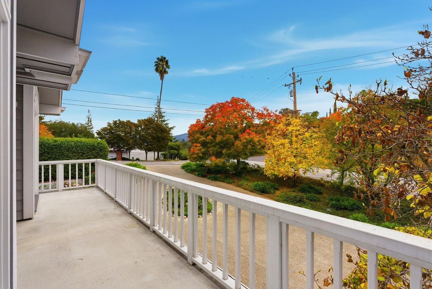 This image showcases a charming balcony with a white railing, offering a serene view of the surrounding landscape. The scene features vibrant autumn foliage, a palm tree, and a glimpse of the road, creating a picturesque and inviting outdoor space. The balcony's neutral tones complement the natural beauty, making it an ideal spot for relaxation and enjoying the scenery.
