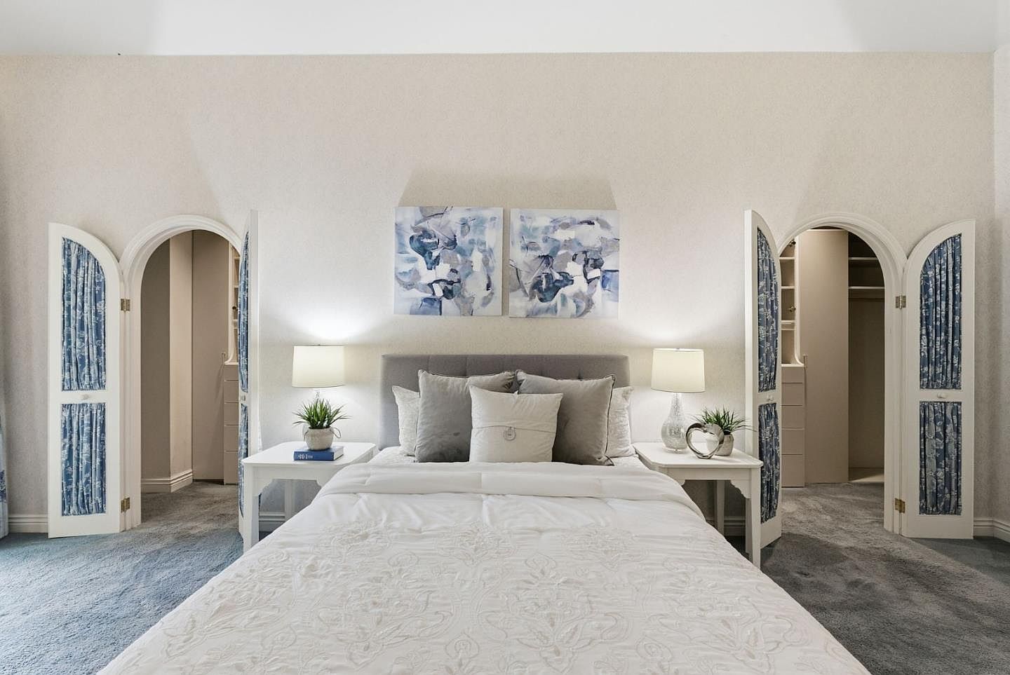 This is a well-lit primary bedroom featuring a bed with white bedding and several pillows, flanked by matching white nightstands with lamps and decorative plants. Two abstract paintings hang above the headboard, and arched doorways with blue and white patterned curtains lead to walk-in closets on either side. The room is carpeted in a neutral tone, creating a serene and inviting atmosphere.