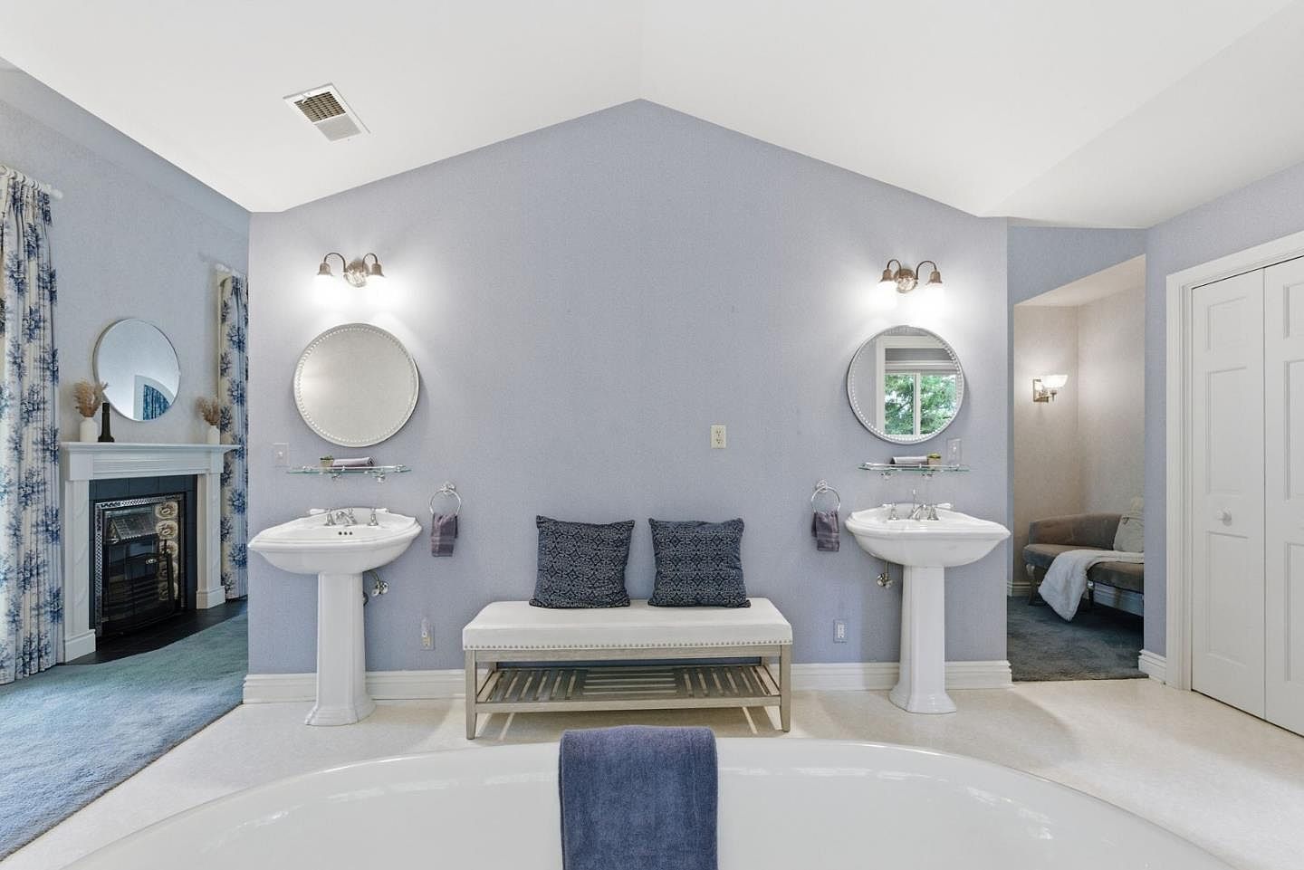This is a well-lit primary bathroom featuring dual pedestal sinks with round mirrors and sconces above. A bench with decorative pillows sits between the sinks, and a large soaking tub is in the foreground. The walls are a soft blue, and the overall style is elegant and serene.