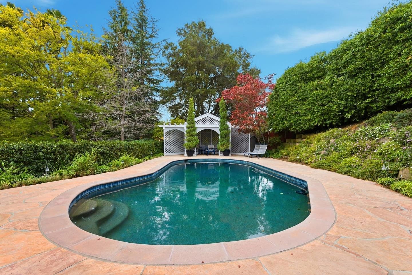 This image showcases a kidney-shaped swimming pool surrounded by a stone patio and lush greenery. A white gazebo with lattice detailing sits at the far end of the pool, adding a touch of elegance. The pool's turquoise water reflects the sky, creating a serene and inviting atmosphere, perfect for relaxation and outdoor entertainment.