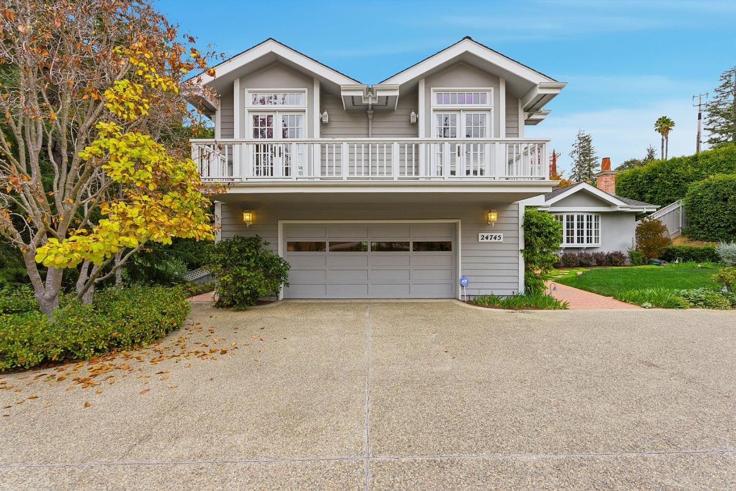 This is a front view of a two-story gray house with a two-car garage. The house features a balcony with white railings above the garage. A well-maintained lawn and landscaping add to the curb appeal, and the driveway is spacious.