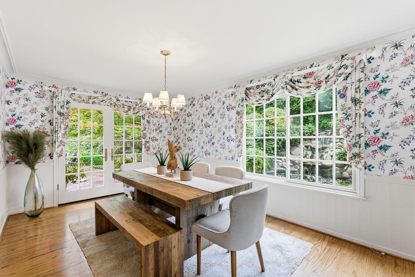 This dining room features a rustic wooden table with a bench and chairs, set on a light-colored rug. The walls are adorned with floral wallpaper, complemented by matching valances over the windows and doors, which provide natural light and views of the greenery outside. A chandelier hangs above the table, adding a touch of elegance to the room's design.