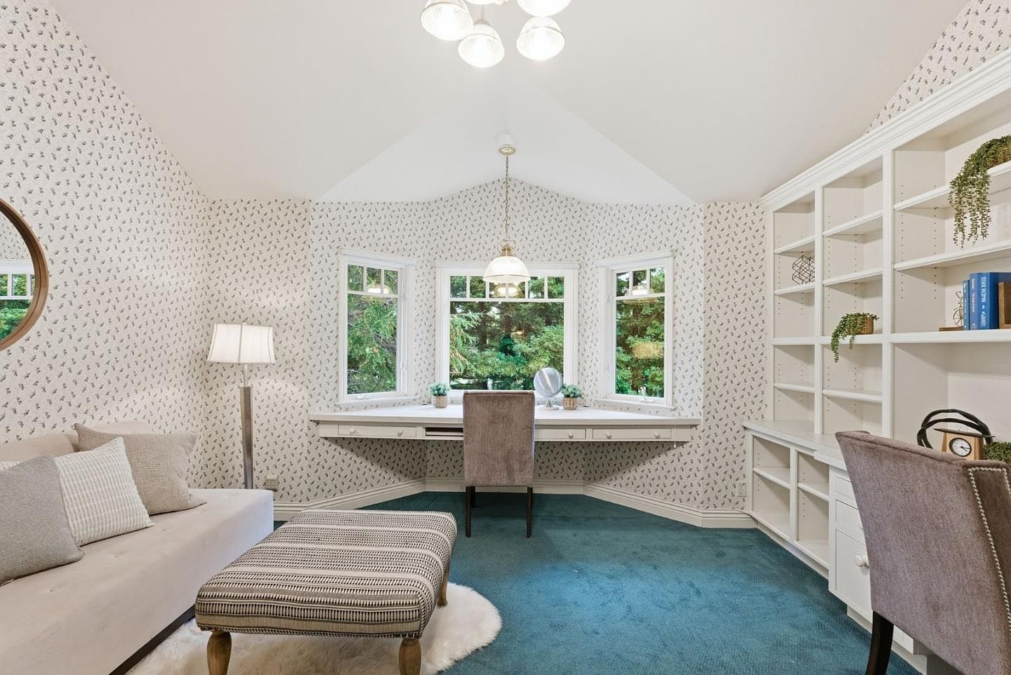 This is an interior shot of a charming home office or study. The room features a built-in desk beneath a bay window, flanked by a built-in bookcase. The room is decorated with patterned wallpaper and has a cozy seating area with a sofa and ottoman, creating a comfortable and functional workspace.