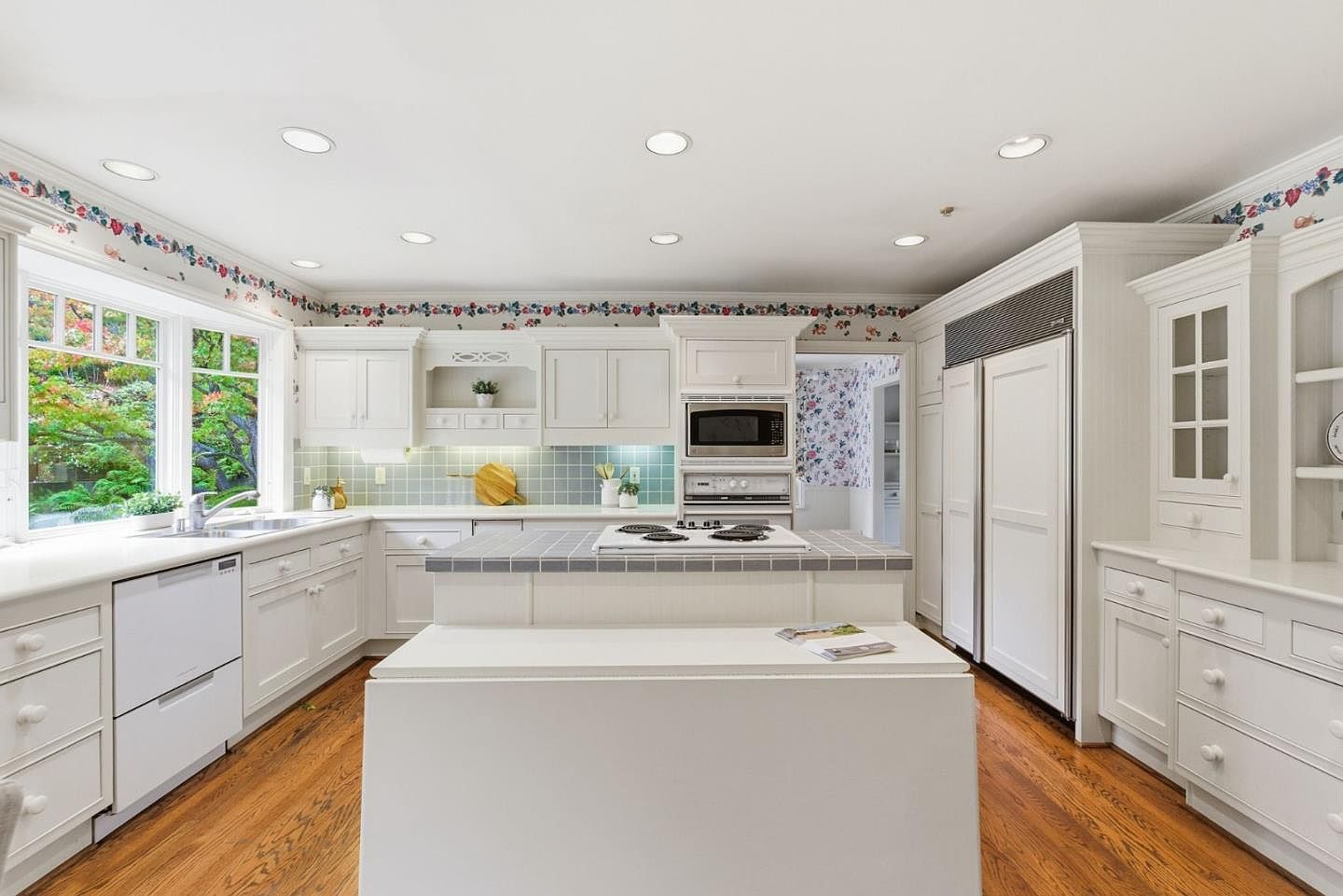 This is a bright and spacious kitchen featuring white cabinetry, a central island with a cooktop, and stainless steel appliances. A large bay window provides ample natural light and a view of the outdoors. The kitchen has a classic, well-maintained appearance with hardwood floors and decorative wallpaper trim.