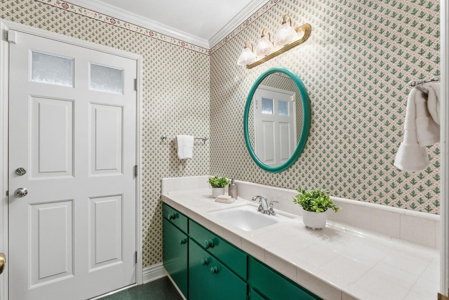 This is a bathroom featuring a white sink and countertop with green cabinets underneath. The walls are covered in a patterned wallpaper, and an oval mirror hangs above the sink. A white door with glass panels is visible to the left, and a towel rack with a white towel is mounted on the wall.