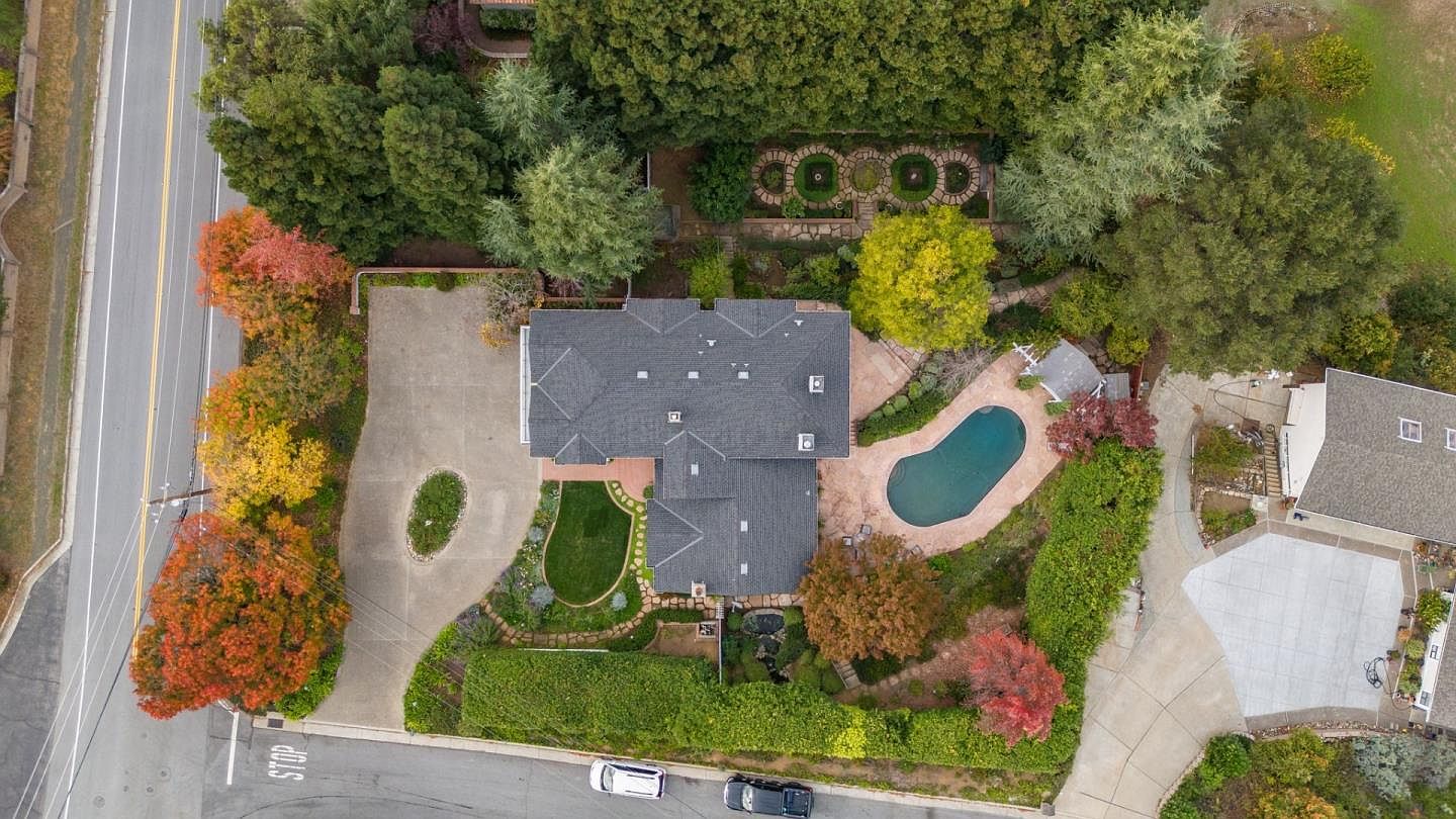 This aerial shot showcases a beautifully landscaped property featuring a house with a dark gray roof, a swimming pool, and meticulously maintained gardens. The driveway leads to the house, and mature trees surround the property, providing privacy and a sense of seclusion. The image highlights the property's curb appeal and outdoor living spaces.