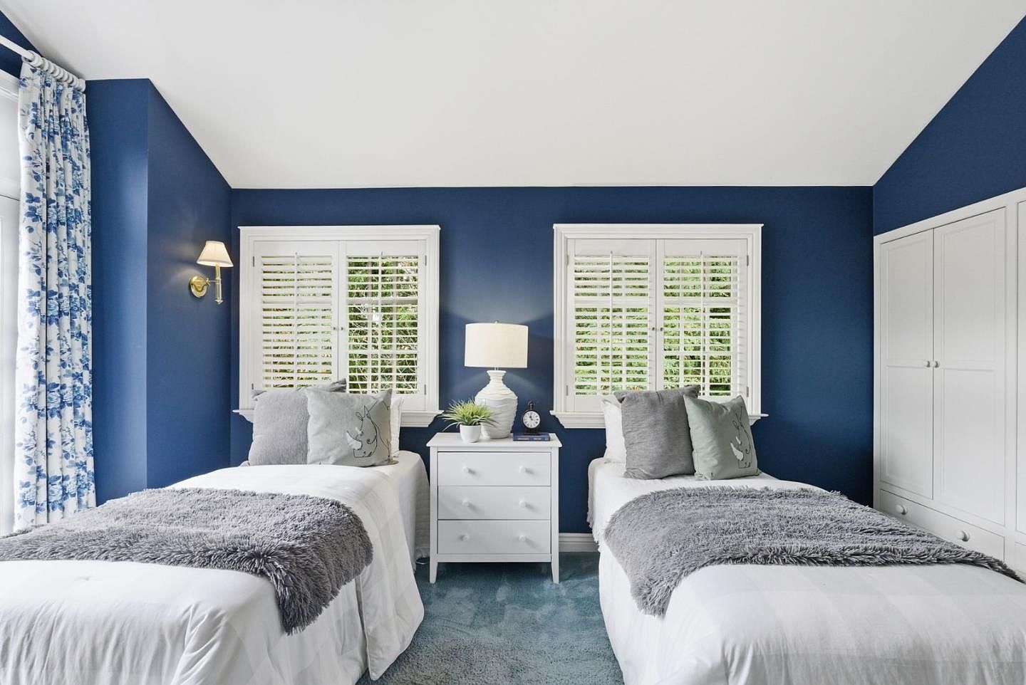 This is a well-lit bedroom featuring two twin beds with white bedding and gray throws. A white nightstand sits between the beds, topped with a lamp and decorative items. The walls are painted a deep blue, and white shutters cover the windows, creating a cozy and inviting atmosphere.