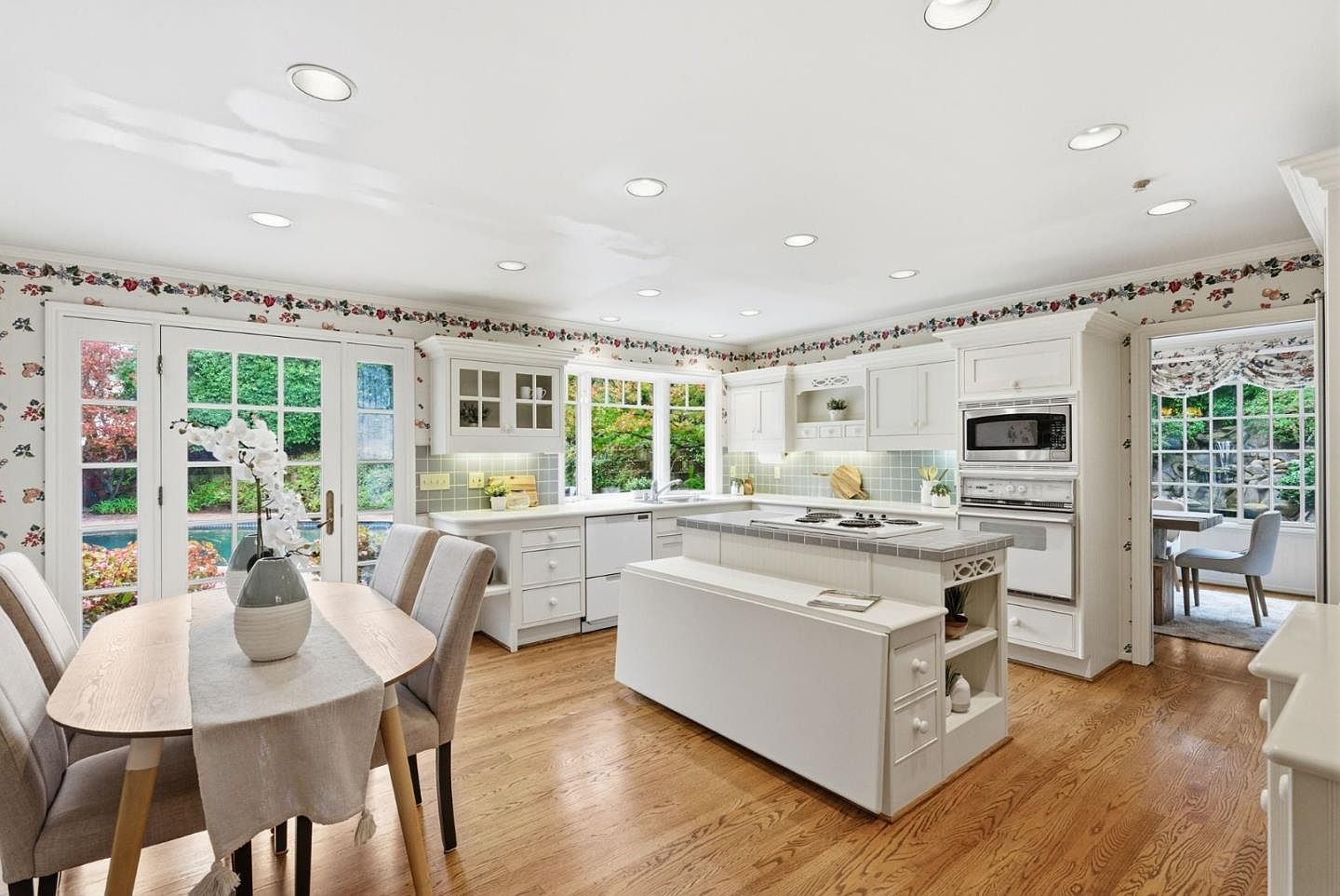 This is a bright and airy kitchen featuring white cabinetry, a central island with a cooktop, and hardwood flooring. The kitchen includes a dining area with a table and chairs, and large windows and doors provide ample natural light and views of the outdoors. The overall style is traditional and well-maintained, creating a welcoming atmosphere.
