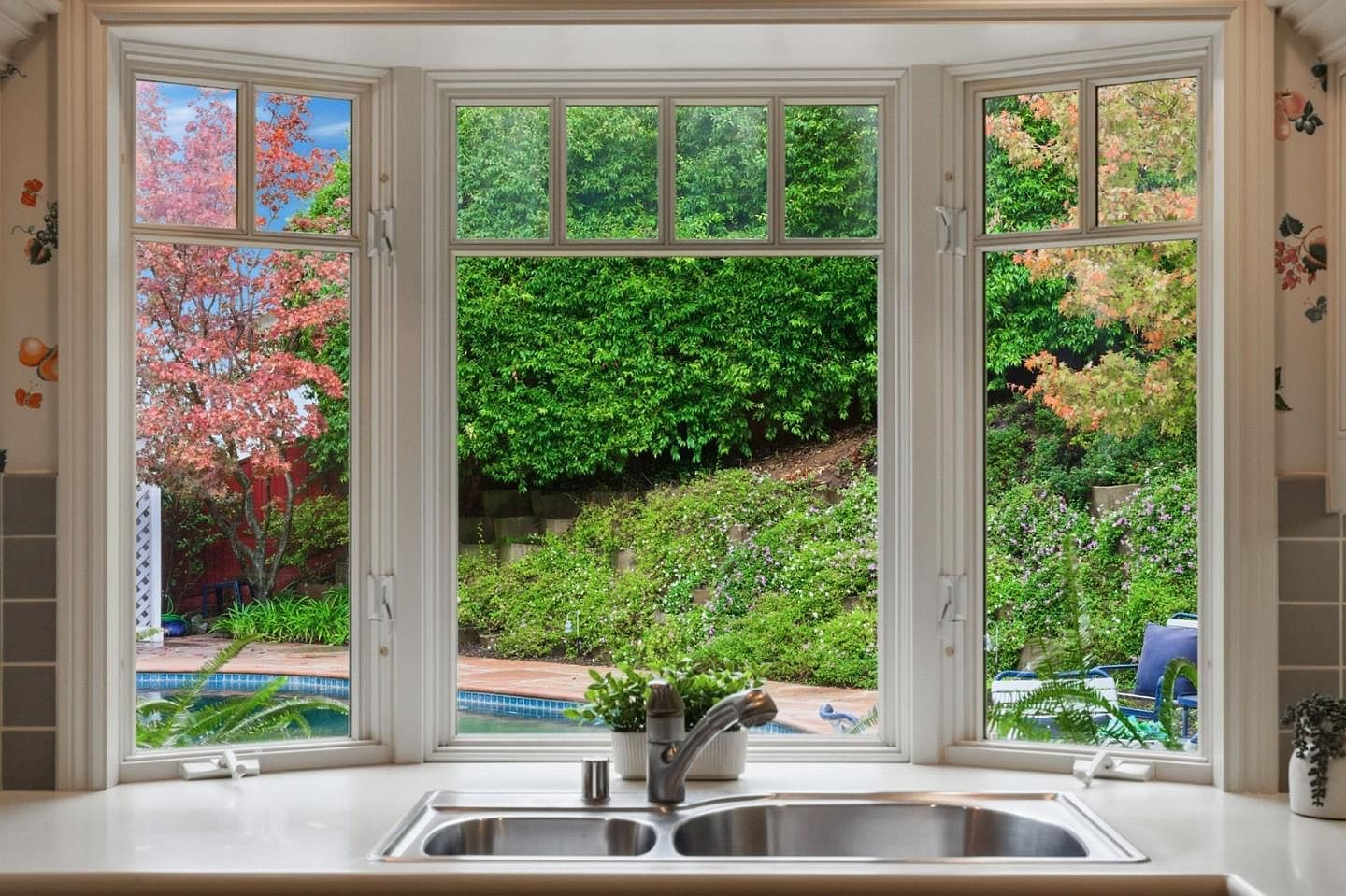 This image showcases a kitchen sink area with a large bay window offering a view of a lush garden and a glimpse of a swimming pool. The window is framed in white, complementing the light-colored countertop and creating a bright, airy atmosphere. The scene provides a sense of tranquility and connection to the outdoors, enhancing the kitchen's appeal.