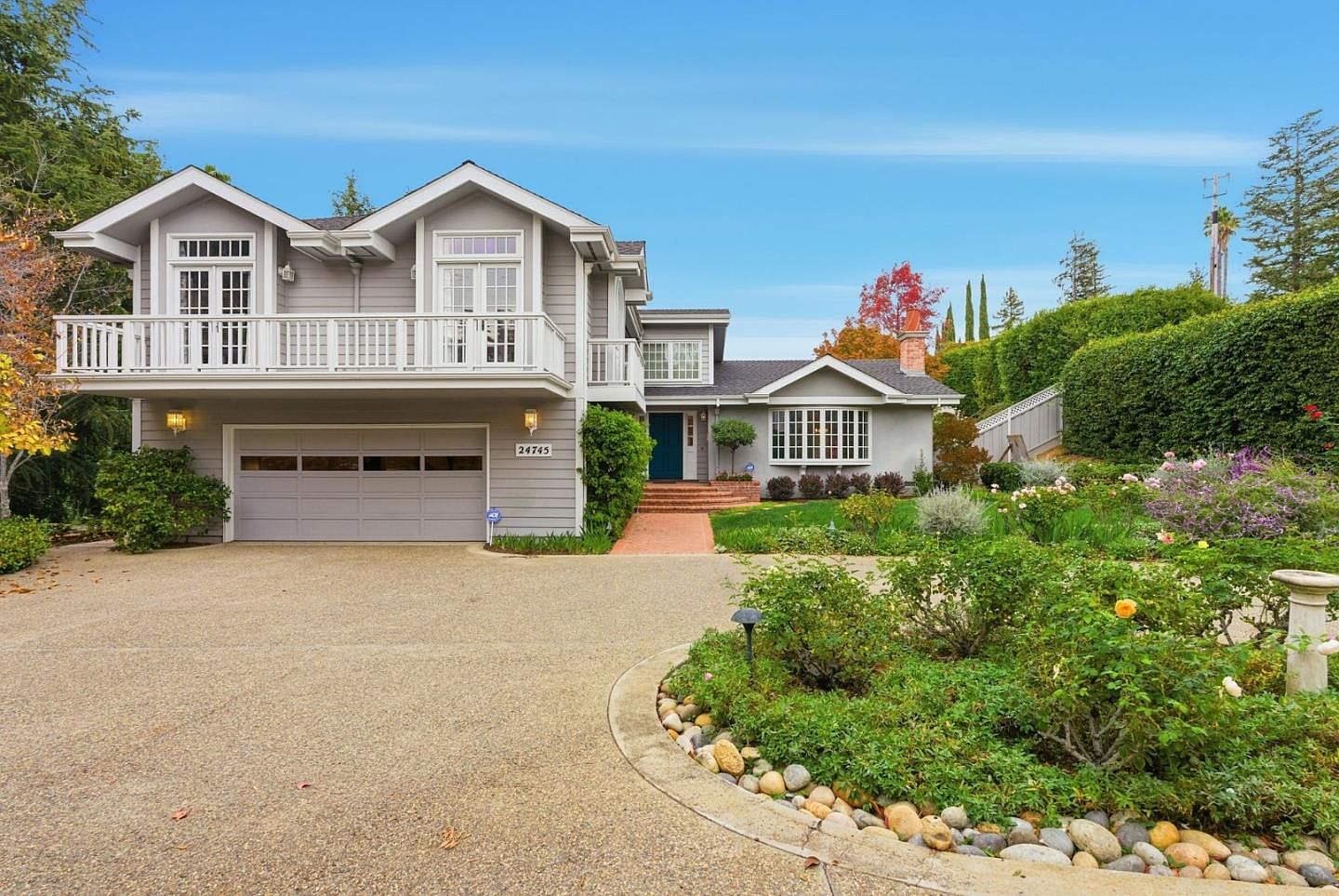 This is a front view of a two-story house with a gray exterior and white trim. The house features a balcony above the garage and a well-maintained front yard with a variety of plants and flowers. A brick pathway leads to the front door, and the driveway is made of gravel.