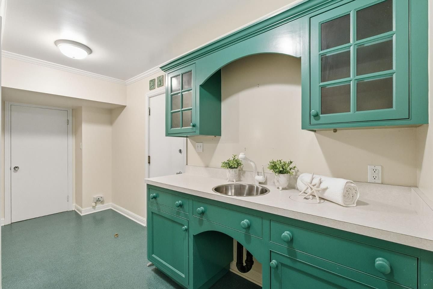 This laundry room features custom green cabinetry with glass-fronted upper cabinets and a built-in sink. The countertops are light-colored, and the room is well-lit with a ceiling light. The floor is a matching green color, creating a cohesive and functional space.