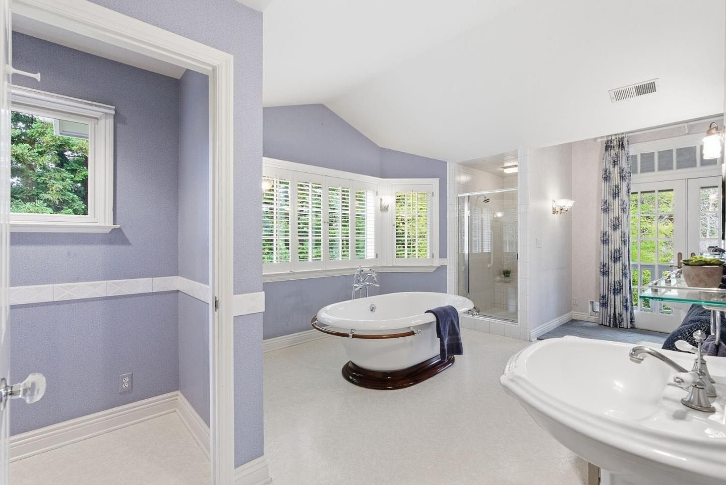 This is a primary bathroom featuring a freestanding bathtub with wood trim, a separate glass-enclosed shower, and a pedestal sink. The walls are covered in a light blue patterned wallpaper, and natural light streams in through shuttered windows. The overall style is elegant and traditional.