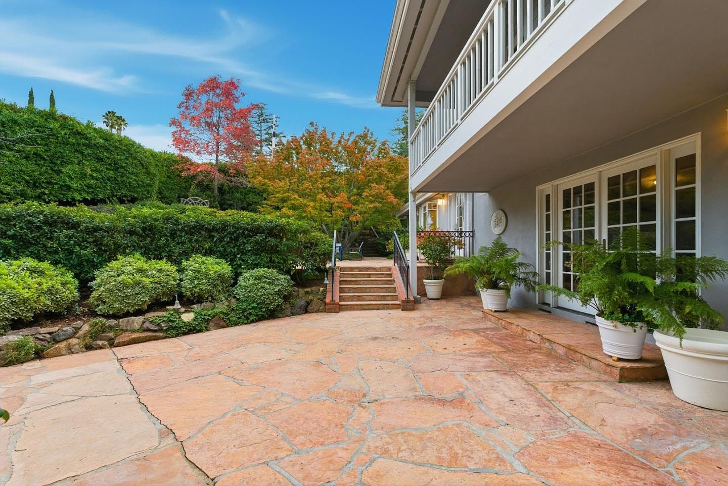 This image showcases a spacious patio area with flagstone paving, leading to a balcony and stairs. Lush greenery and trees surround the patio, creating a serene and private outdoor space. The architectural style of the house is traditional, with white railings and trim, offering a blend of elegance and natural beauty.