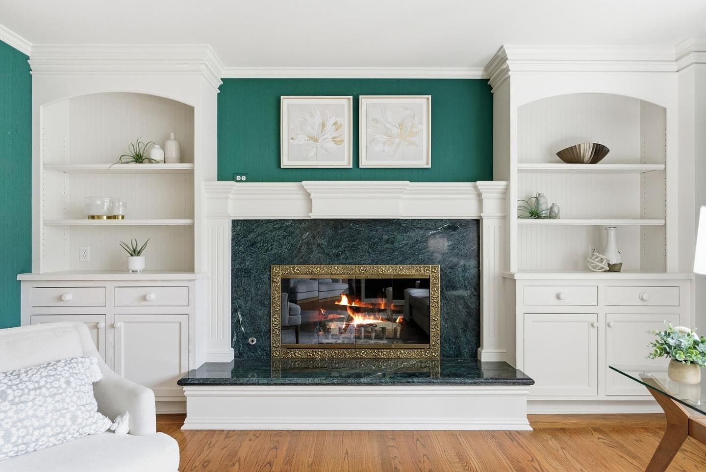 This interior shot showcases a living room with a prominent fireplace flanked by built-in shelving units. The fireplace features a green marble surround and a decorative gold screen, adding a touch of elegance. The shelves are styled with decorative items, and the room is painted in a rich teal color, creating a cozy and inviting atmosphere.