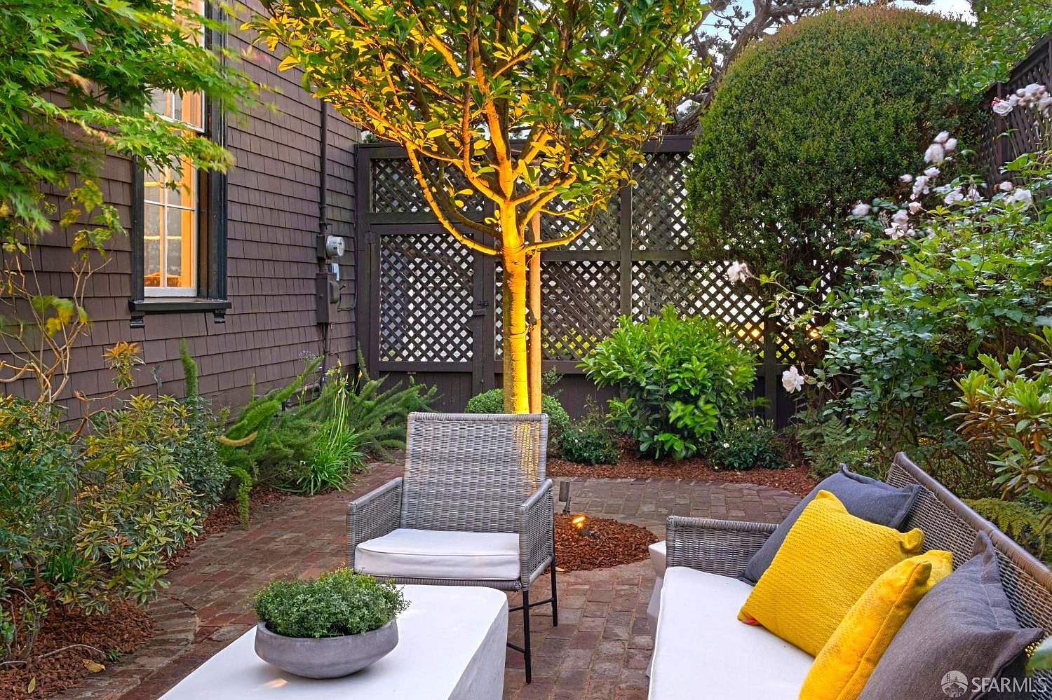This inviting outdoor patio space features a brick-paved floor and comfortable modern wicker-style seating with plush cushions and vibrant yellow accent pillows. A central tree is illuminated by warm uplighting, creating a cozy atmosphere against the backdrop of a dark-stained lattice privacy fence. The scene is framed by lush greenery and a side view of the home's dark shingled exterior, offering a serene and private retreat.