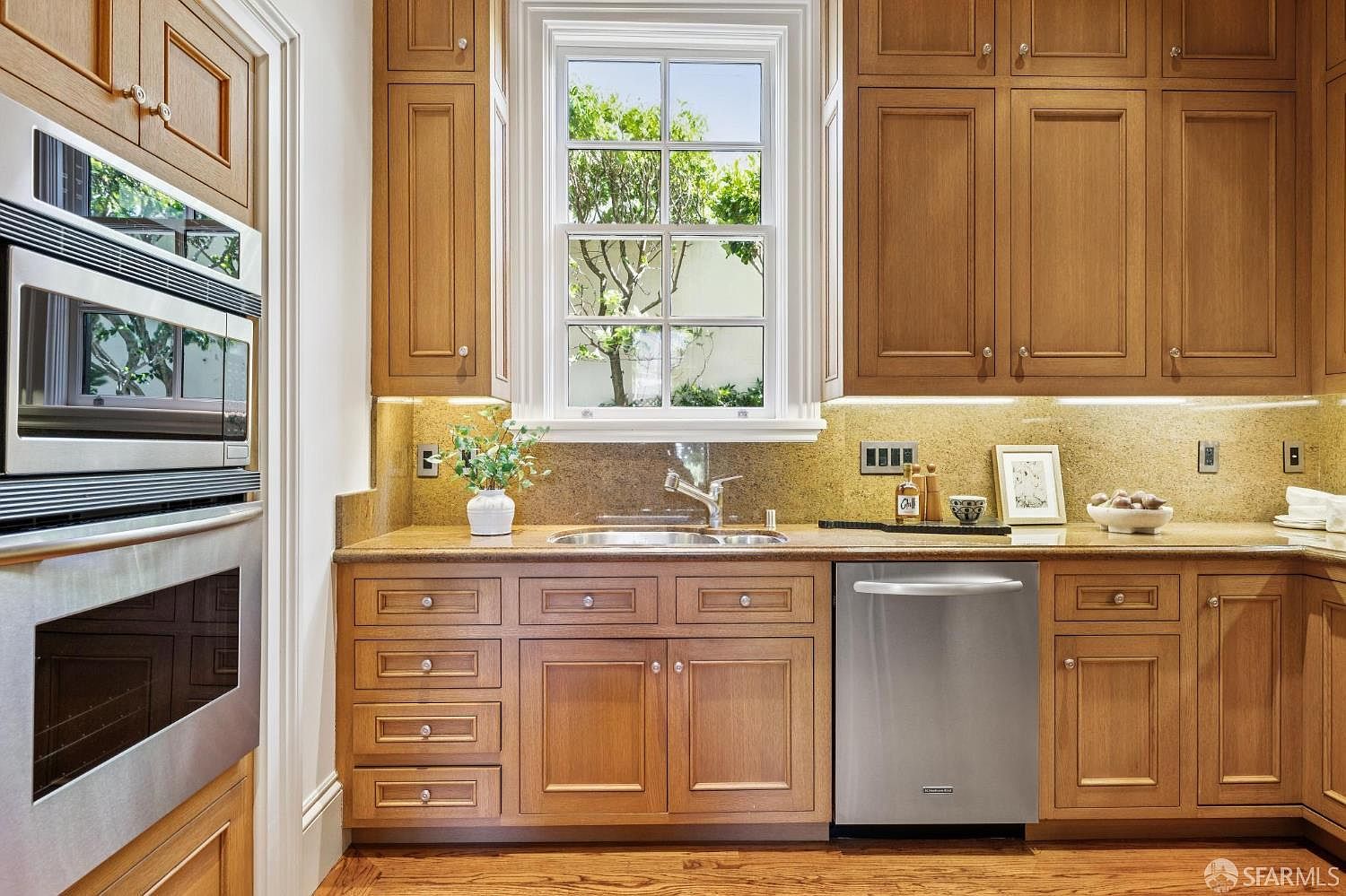 This well-appointed kitchen features warm-toned wood cabinetry, a stainless steel dishwasher, and a built-in microwave oven. A central window provides natural light over the sink, while stone countertops and a matching backsplash create a cohesive look. The space is organized and functional, showcasing a classic design with ample storage and a clean, polished aesthetic.