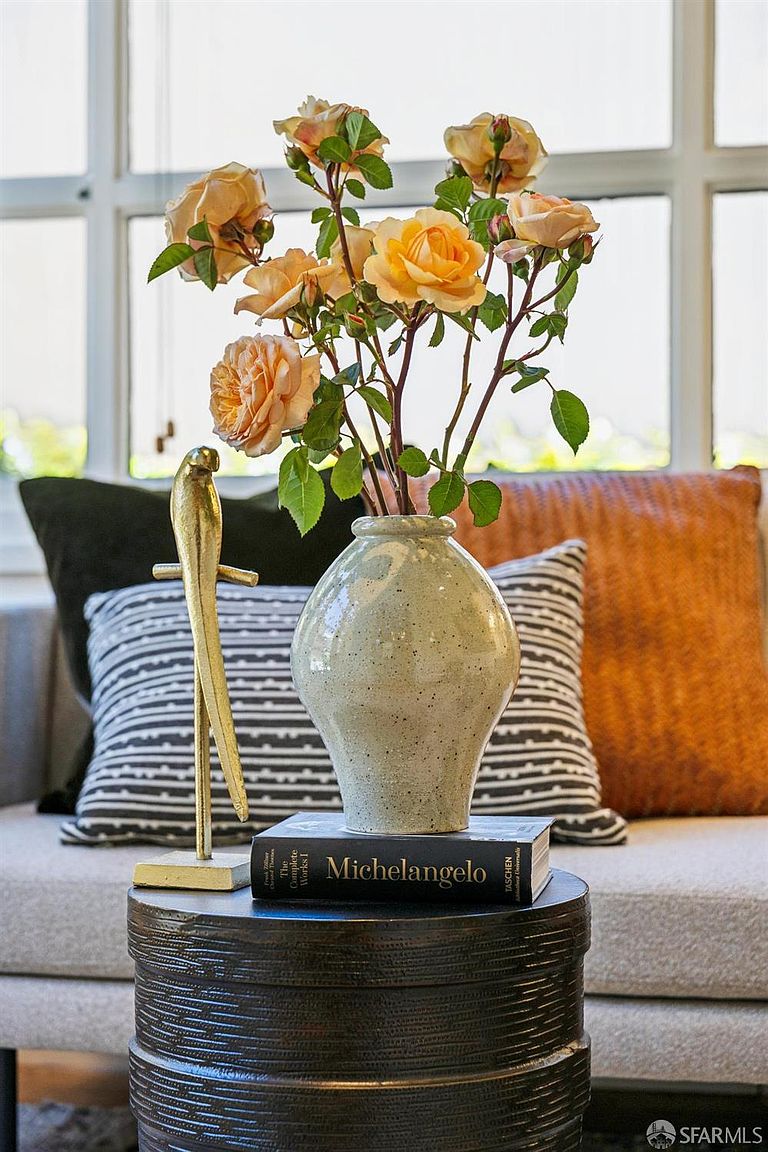 This close-up shot captures a sophisticated living room vignette featuring a textured, dark cylindrical side table topped with a ceramic vase of peach-colored roses. A gold bird figurine stands beside the vase, which rests on a book titled 'Michelangelo'. In the background, a neutral-toned sofa is accented with a black-and-white striped pillow and a solid orange pillow, creating a warm and inviting atmosphere.