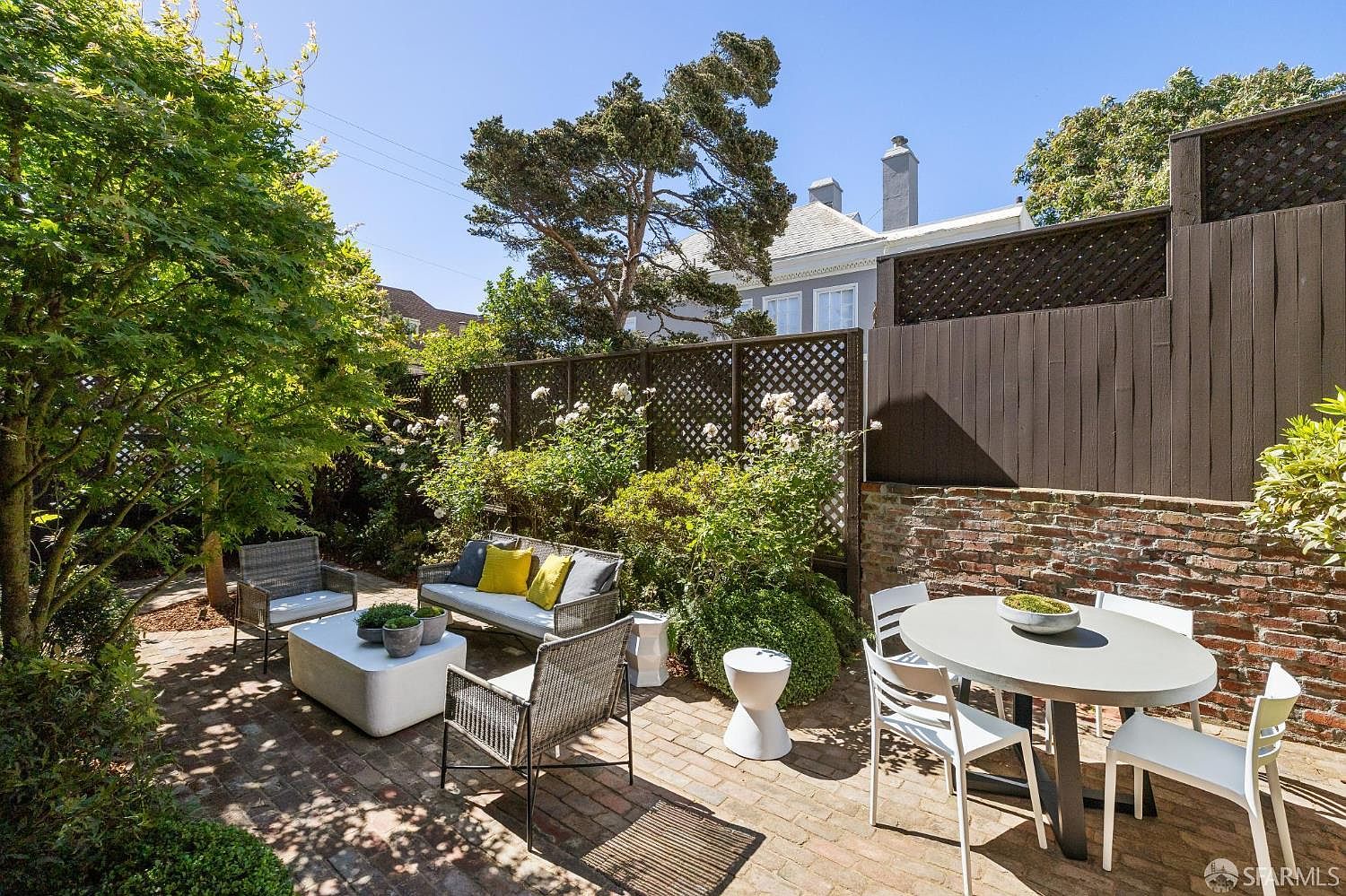 This inviting outdoor patio space features a classic brick-paved floor and is furnished with a comfortable seating area and a separate dining set. The area is enclosed by a decorative lattice fence and a rustic brick wall, creating a private and serene garden atmosphere. Lush greenery, including a mature tree and flowering bushes, frames the space, offering a peaceful retreat for outdoor relaxation.