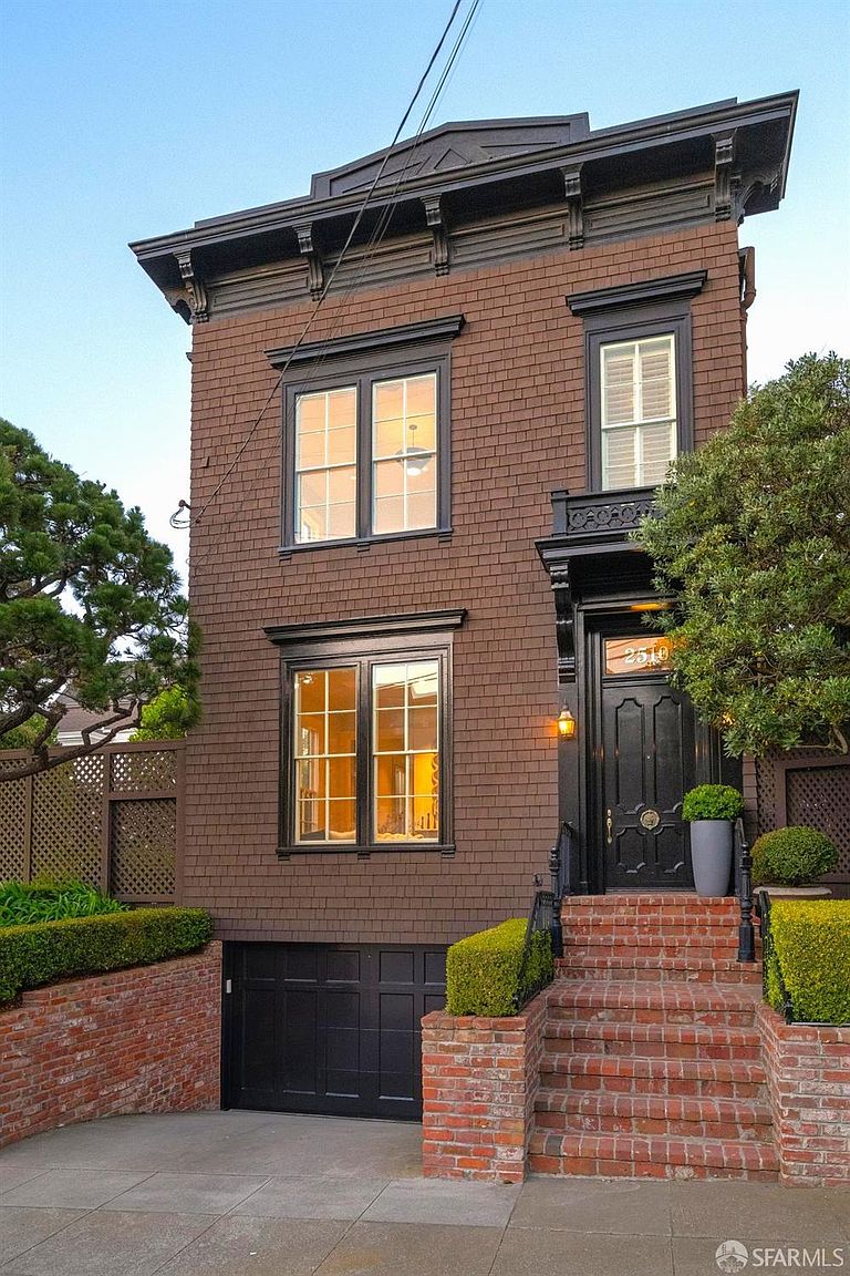 This charming, multi-story residence features a dark brown shingled facade with elegant black trim and a prominent, ornate cornice. A brick staircase leads up to a sophisticated black front door, while a lower-level garage is integrated into the foundation. The property is framed by manicured hedges and a mature tree, presenting a classic and well-maintained urban aesthetic.