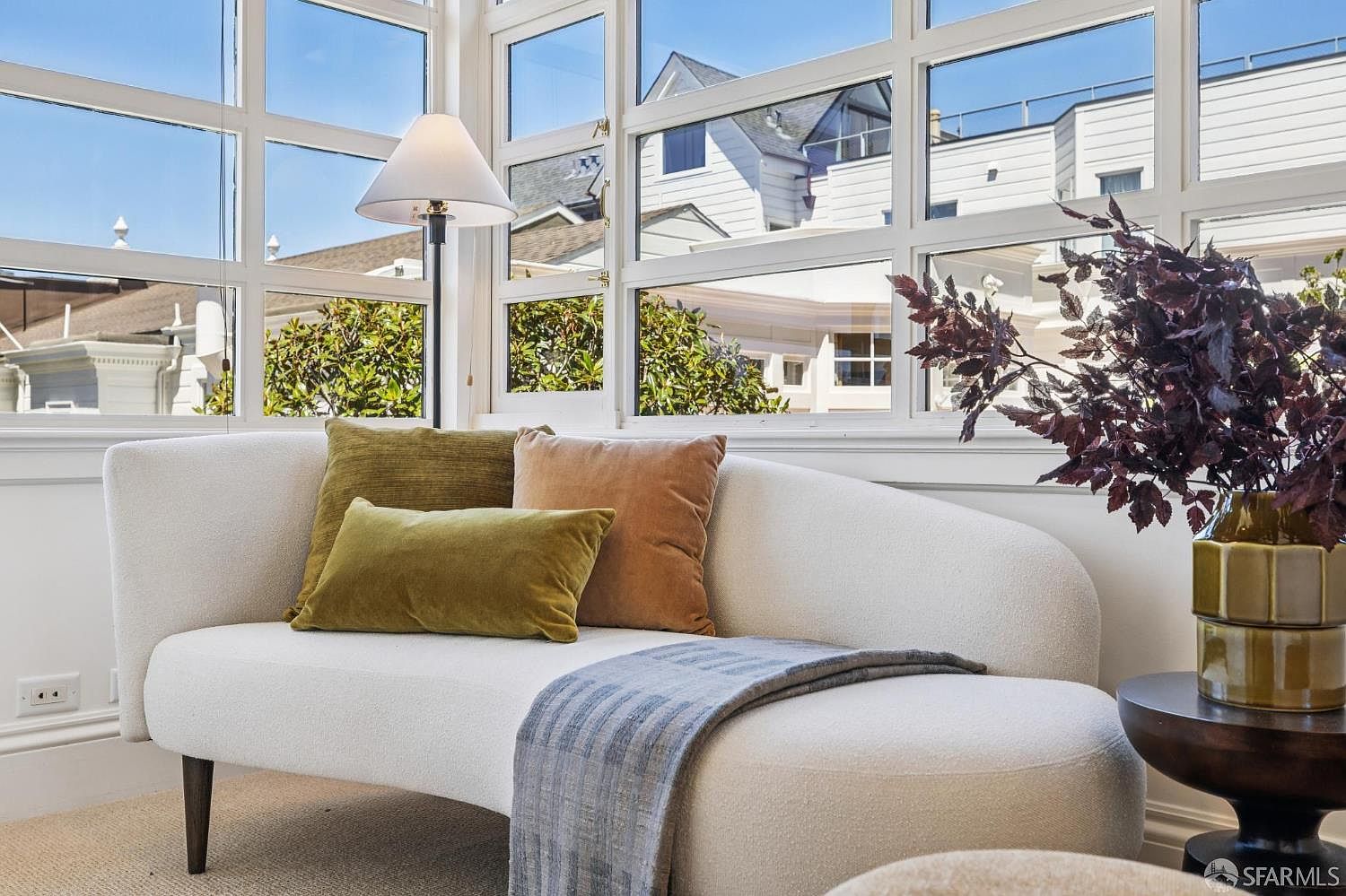 This bright and inviting sunroom features a curved, cream-colored settee adorned with olive and rust-toned throw pillows. A tall floor lamp stands beside the seating area, which is positioned in front of large, multi-pane windows that offer a view of neighboring rooftops. A small side table holds a decorative vase with dark foliage, creating a cozy and sophisticated reading nook.