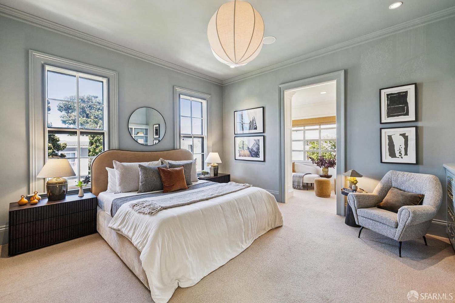 This elegant primary bedroom features a soft, neutral color palette with light blue-gray walls and plush carpeting. The room is anchored by a curved, upholstered headboard and a large, textured pendant light fixture that serves as a central focal point. A cozy seating area with a gray armchair and a doorway leading to an adjacent sunlit space enhance the room's sophisticated and tranquil atmosphere.