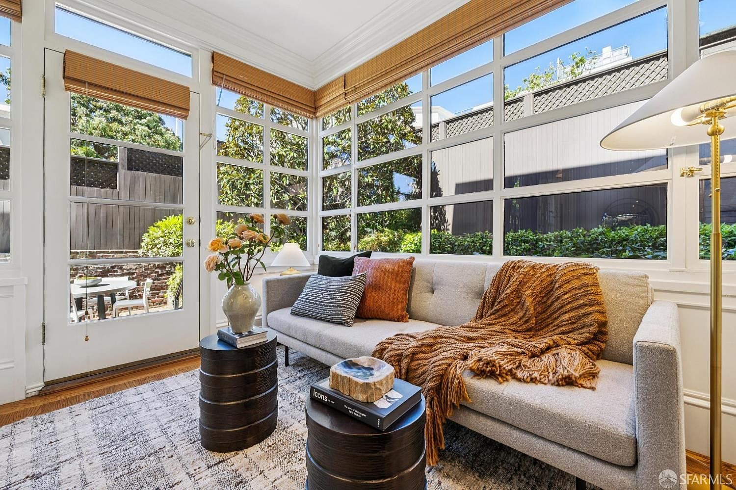 This bright sunroom features a light gray sofa adorned with textured throw pillows and a warm, chunky knit blanket. The space is framed by large windows with wooden blinds that offer a clear view of the outdoor patio and greenery, creating a seamless connection to the garden. Two dark, cylindrical side tables serve as focal points, one holding a vase of flowers and the other a decorative bowl, enhancing the cozy and inviting atmosphere.