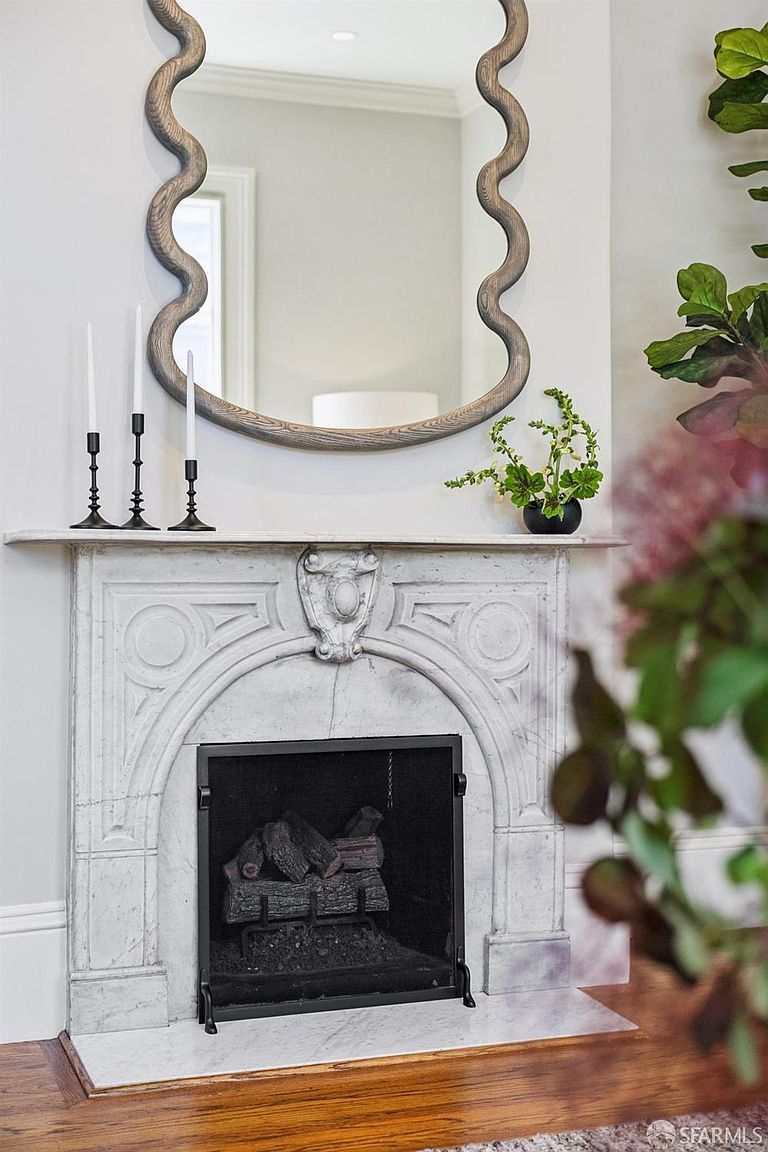This elegant living room feature centers on a classic stone fireplace with an ornate, carved mantel and a black metal fire screen. Above the mantel, a large, wavy-edged mirror reflects the room, flanked by a trio of slender black candlesticks and a small potted plant. The scene is captured from a straight-on perspective, highlighting the sophisticated, traditional aesthetic of the hearth area.