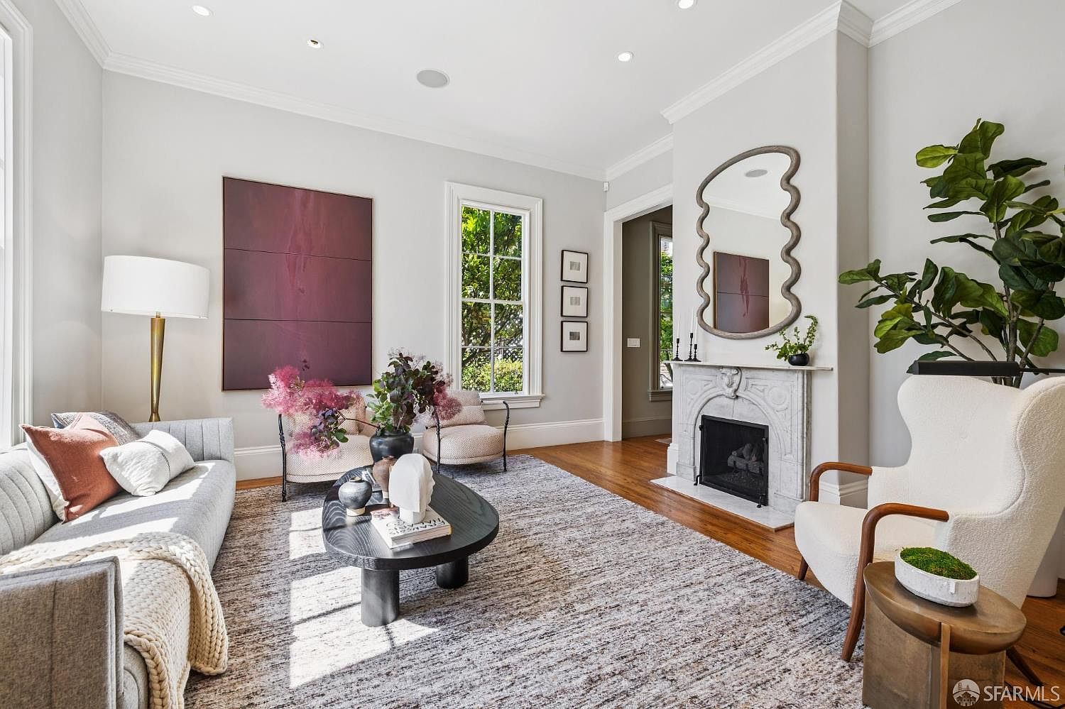 This elegant living room features a sophisticated neutral color palette with a large grey sofa, a textured area rug, and a central black coffee table. A classic fireplace with a detailed stone surround and a wavy-edged mirror serves as a focal point, complemented by a large piece of abstract art on the adjacent wall. The space is brightened by natural light from a window and includes a comfortable white armchair, creating a refined and inviting atmosphere.