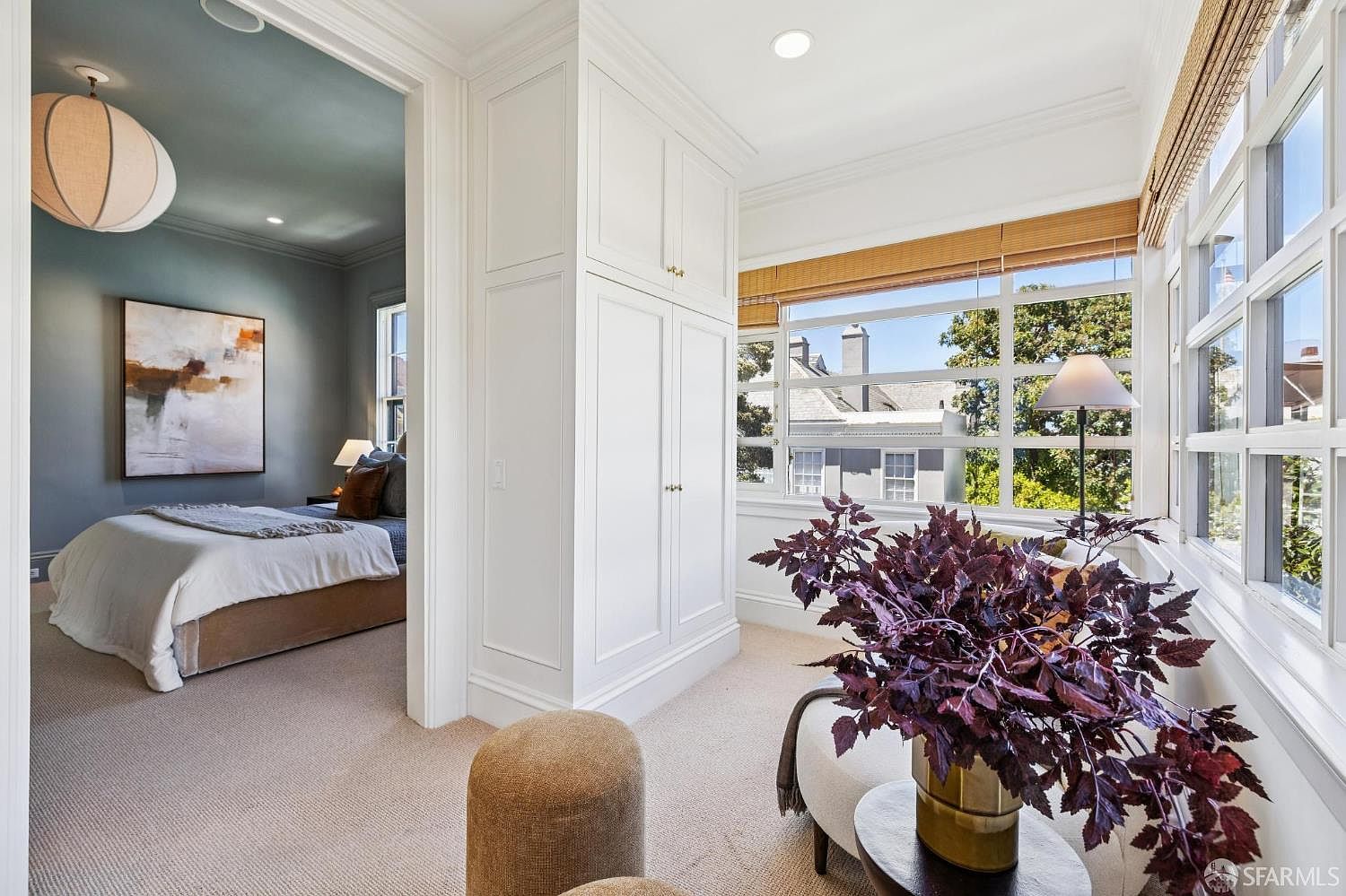 This bright sunroom features a large window wall with woven shades, offering a view of the neighborhood. The space is furnished with a comfortable chair, a small side table holding a potted plant, and two textured ottomans, creating a cozy reading nook. A doorway leads into an adjacent bedroom, which is decorated in soft blue tones and features a large piece of abstract wall art.
