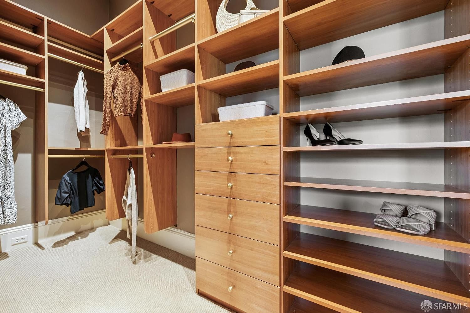 This organized walk-in closet features custom light wood shelving and cabinetry, providing ample storage for clothing and accessories. The layout includes hanging rods, open shelving for shoes and storage bins, and a central stack of six drawers with gold-toned hardware. The space is finished with neutral carpeting, creating a clean and functional storage area.