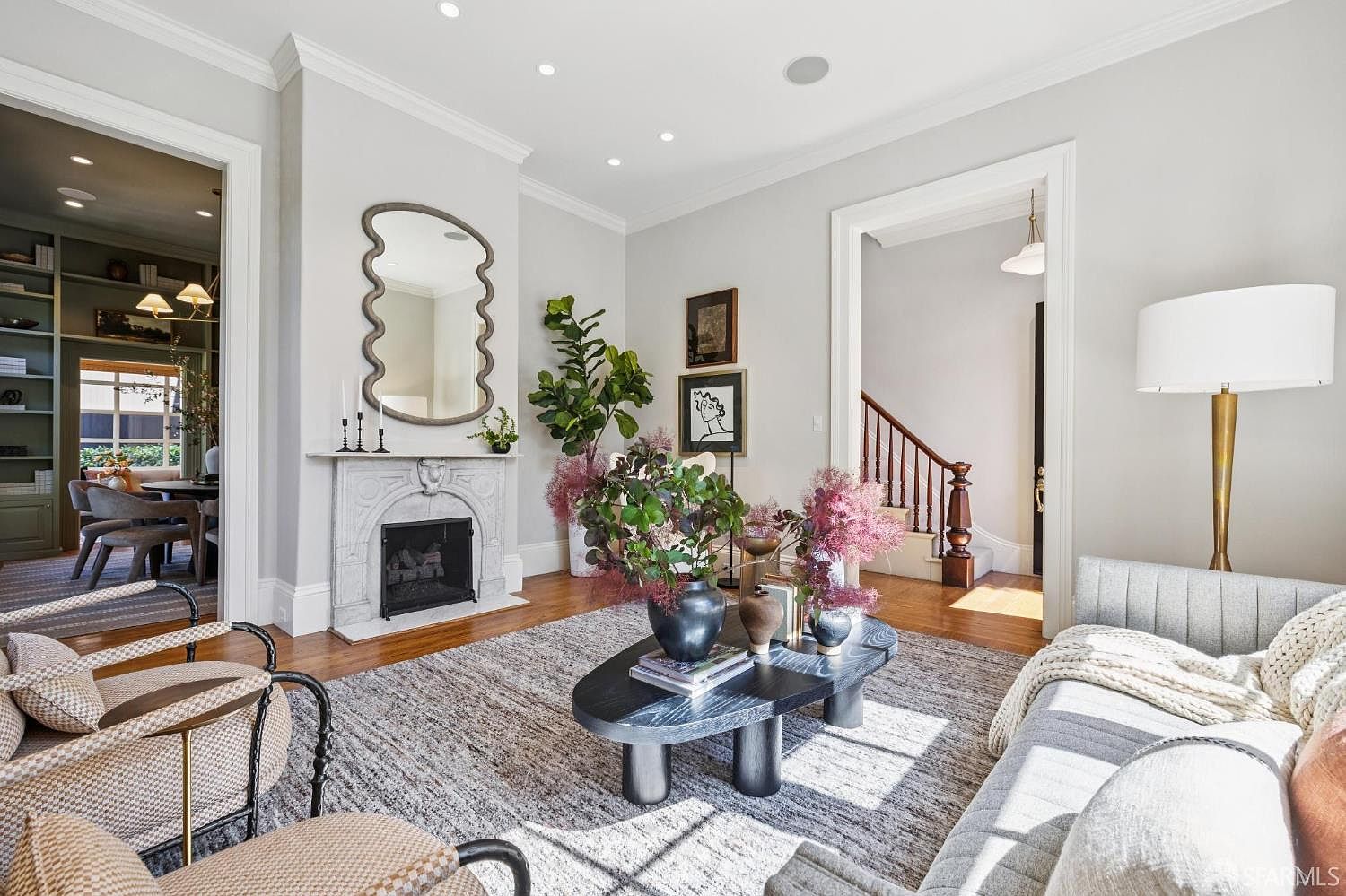 This elegant living room features a sophisticated neutral color palette, centered around a classic fireplace with a decorative stone mantel and a wavy-edged mirror above. The space is furnished with a plush gray sofa, patterned armchairs, and a unique, dark-toned organic-shaped coffee table adorned with floral arrangements. The room flows seamlessly into an adjacent dining area and a hallway with a staircase, creating an open and inviting atmosphere with warm wood flooring and recessed lighting.