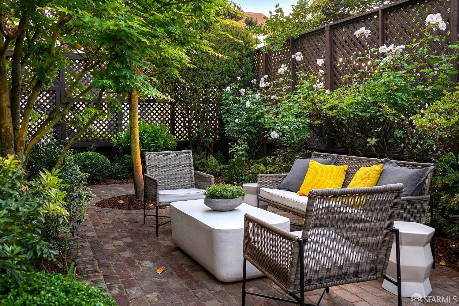 This inviting outdoor patio features a brick-paved floor and a comfortable seating arrangement with a sofa and two armchairs accented by yellow cushions. A low, rectangular white coffee table sits in the center, while a mature tree and lush greenery provide a sense of privacy against the dark lattice fencing. The space is framed by blooming white roses, creating a serene and well-manicured garden atmosphere perfect for relaxation.