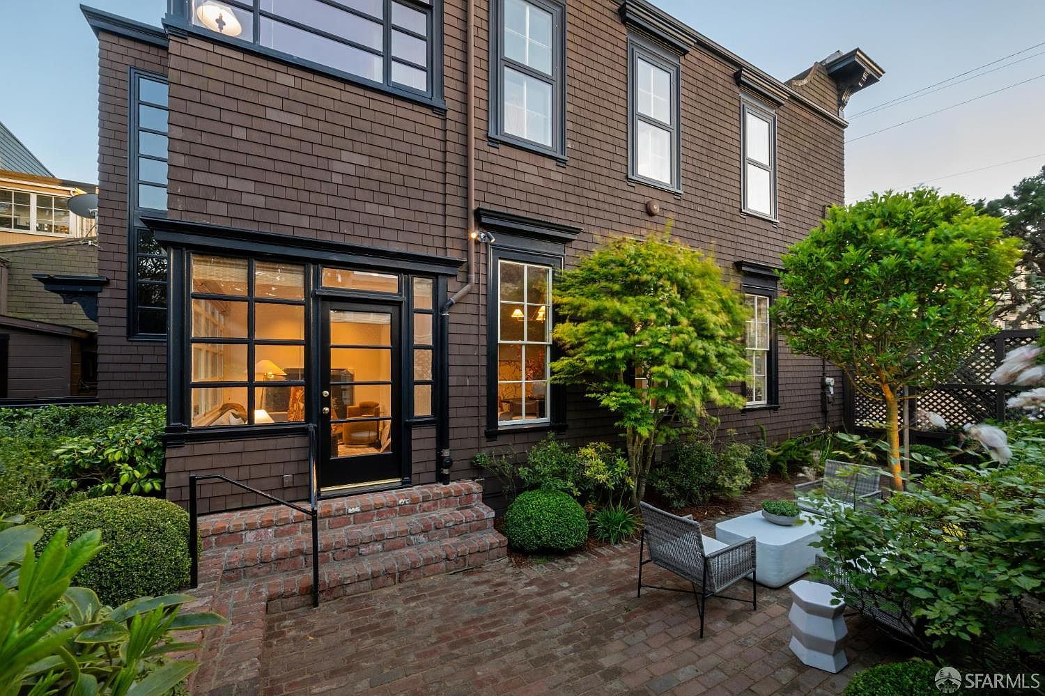 This rear exterior view showcases a multi-story home with dark brown shingle siding and black-trimmed windows. A brick patio area features outdoor seating, surrounded by lush landscaping, including a prominent Japanese maple and manicured shrubs. The perspective is at ground level, capturing the transition between the home's interior living space and the private garden retreat.