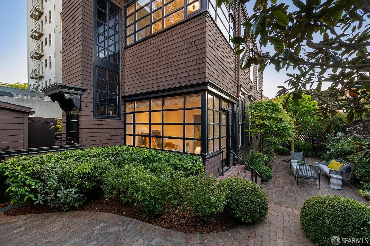 This image captures a charming side yard and patio area featuring a brick-paved terrace and manicured shrubbery. The home's exterior is clad in dark brown shingles and features a prominent, multi-paned glass sunroom extension that provides a view into the interior. The perspective is from the garden looking toward the house, highlighting the blend of architectural detail and outdoor living space.