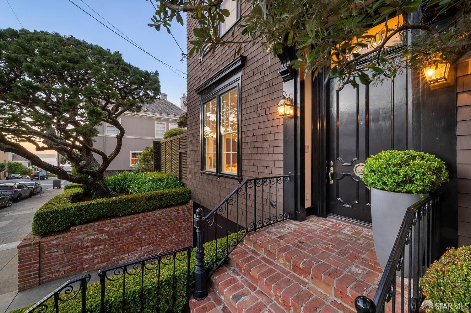 This charming entryway features a classic black front door framed by dark brown shingle siding and elegant black iron railings. A brick staircase leads up to the entrance, which is accented by warm wall-mounted lanterns and a large potted plant. The scene is framed by a mature, sculpted tree and a manicured hedge, creating a sophisticated and welcoming curb appeal.