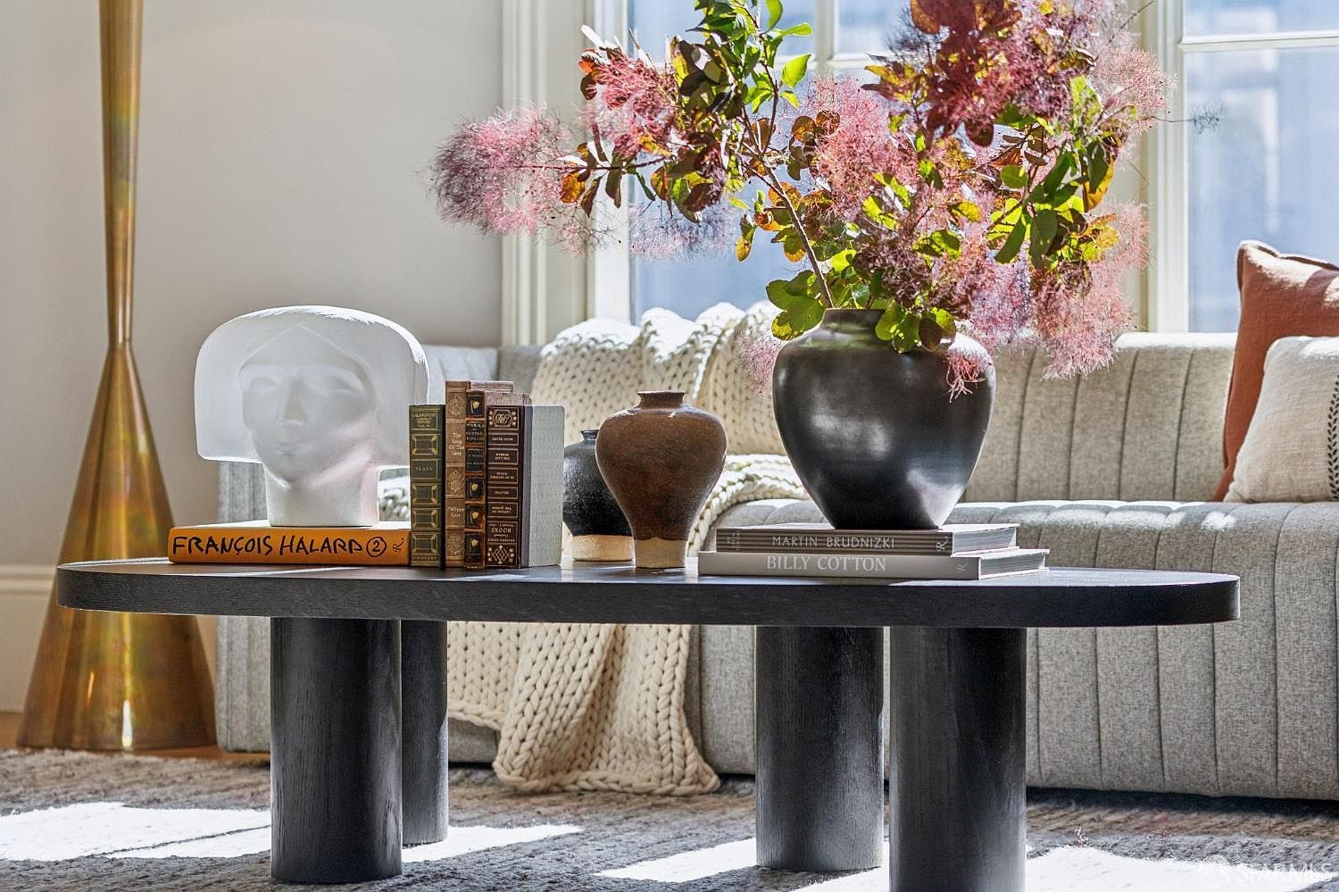 This close-up shot captures a sophisticated living room vignette centered on a dark, cylindrical-legged coffee table. The table is styled with a white sculptural bust, a collection of books, and a dark ceramic vase filled with vibrant, feathery foliage. In the background, a textured, neutral-toned sofa with plush pillows and a draped throw blanket creates a cozy, layered atmosphere bathed in soft natural light.