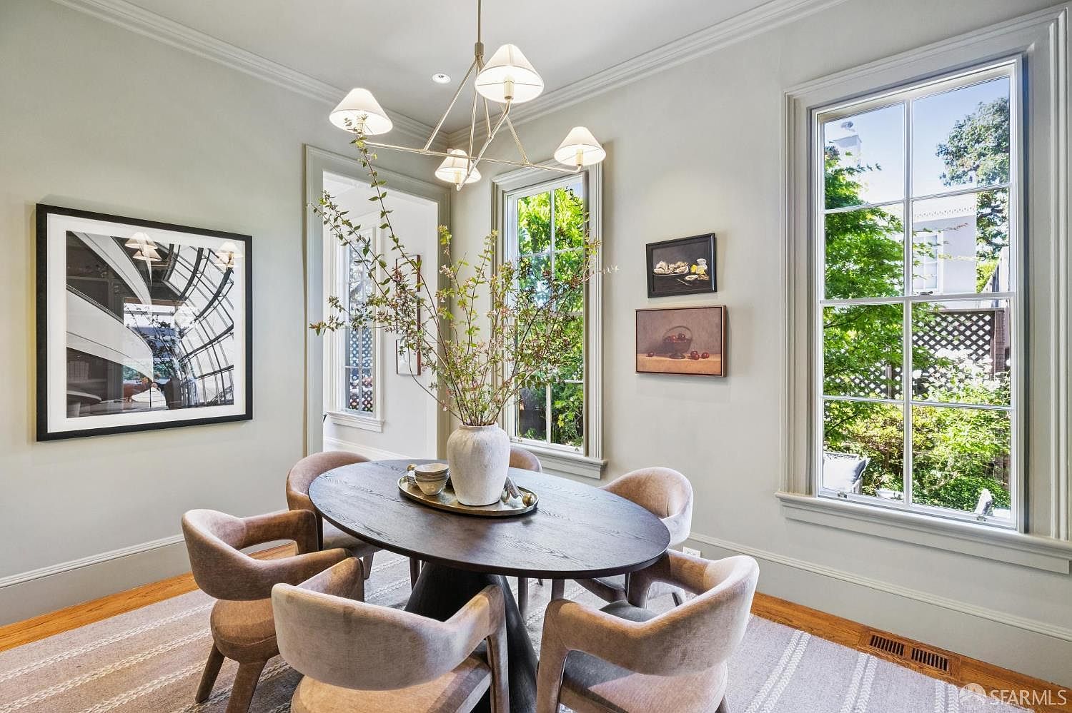 This elegant dining room features a dark oval table surrounded by six upholstered chairs, centered beneath a modern multi-arm chandelier. Large windows on two walls provide ample natural light and views of the lush greenery outside, while neutral wall tones and a textured area rug create a sophisticated, airy atmosphere. The space is accented by framed artwork on the wall and a decorative vase with branches, offering a refined and welcoming setting for meals.